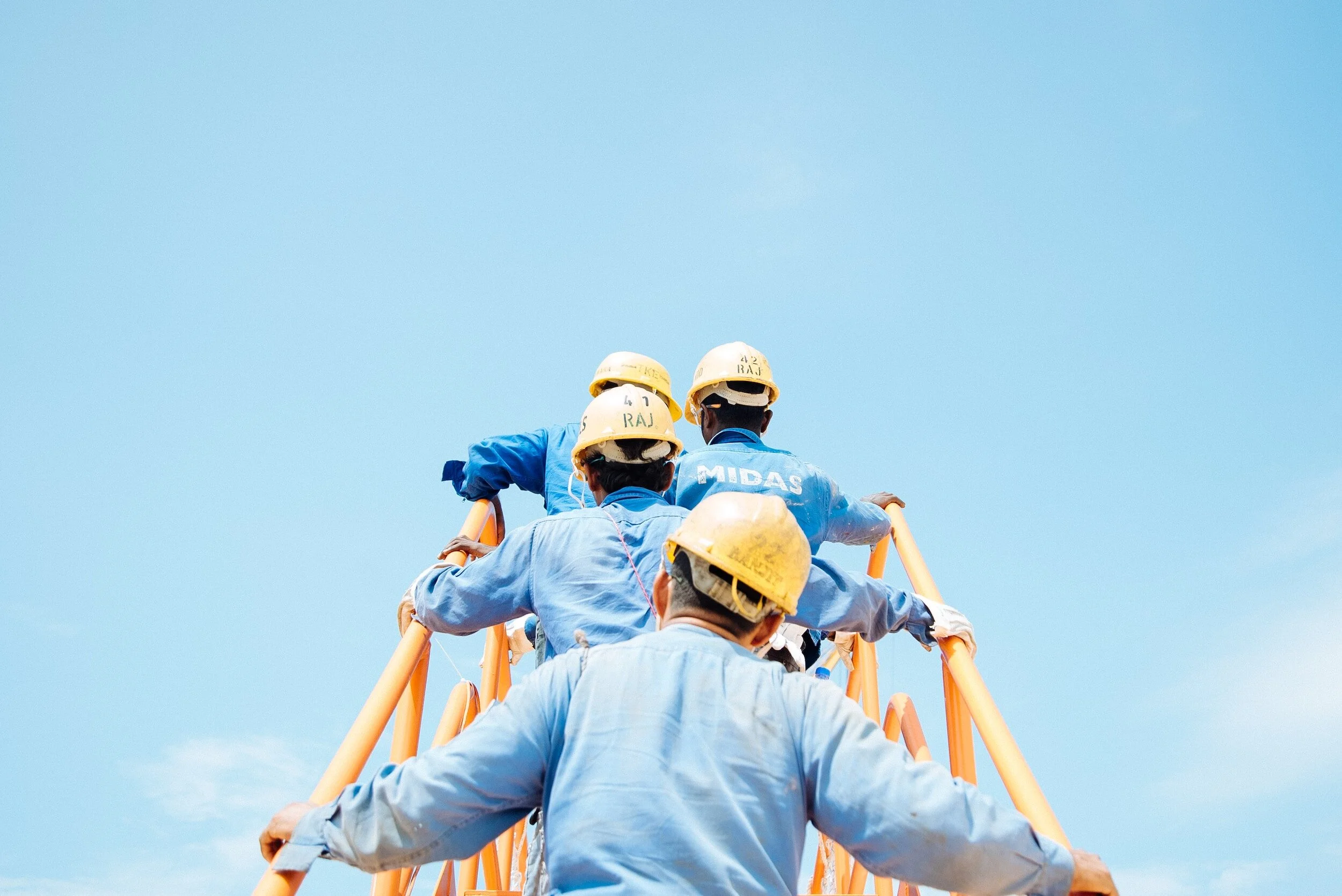 A group of construction workers wearing yellow helmets and blue uniforms are ascending a yellow ladder against a clear blue sky.