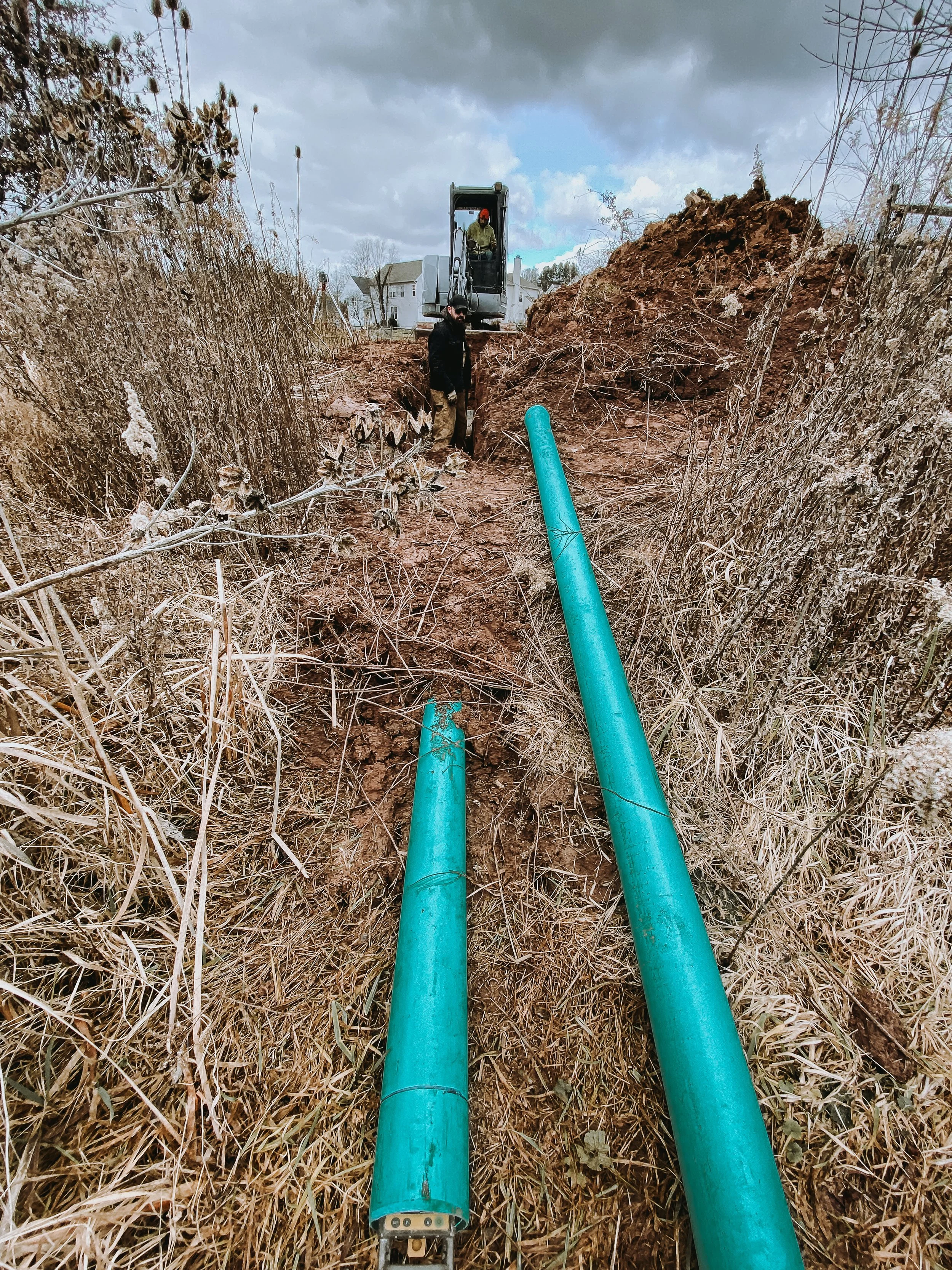 A construction site with two large green pipes laid in the ground, surrounded by dried grass. Two workers are present: one with a black jacket and beard standing near the trench, and another in a construction vehicle operating machinery in the background. Houses and trees are visible in the distance under a cloudy sky.