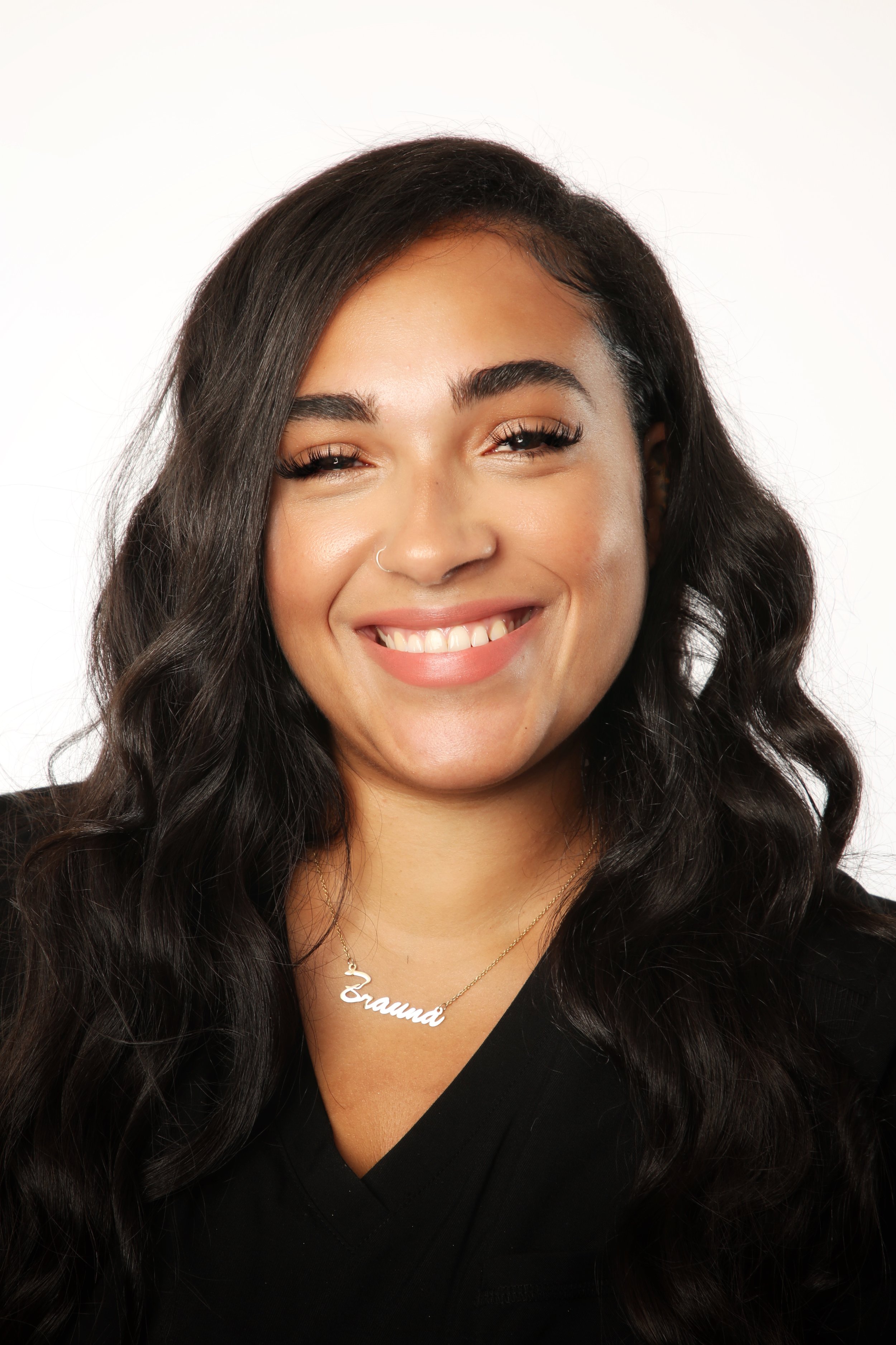 Close-up of a young woman with long, dark curly hair, smiling, wearing a black top and a gold necklace with a nameplate that says 'Brauna' against a plain white background.