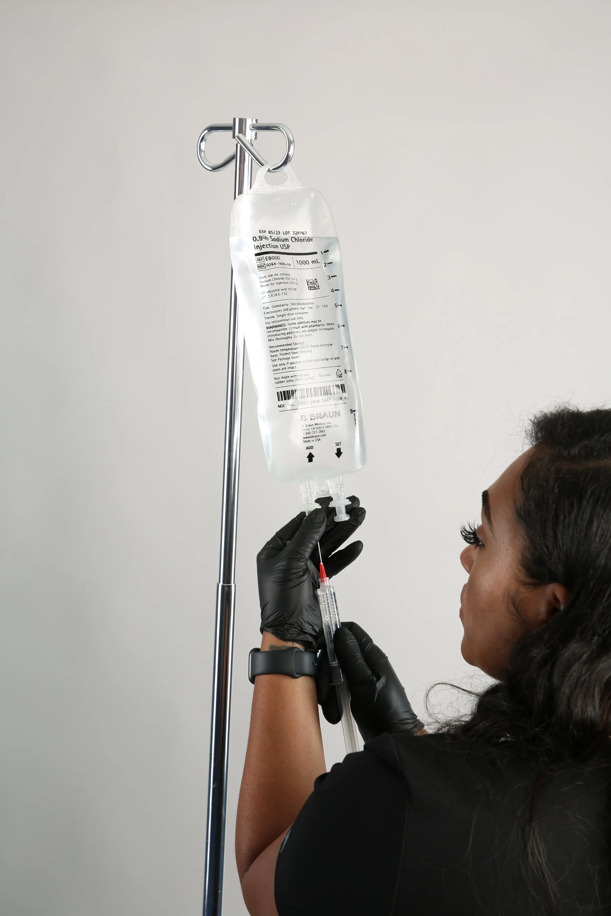 A healthcare worker prepares a syringe with a vaccine needle, next to an IV bag labeled '0.9% Sodium Chloride Injection USP,' hanging on a metal stand, in a clinical setting.