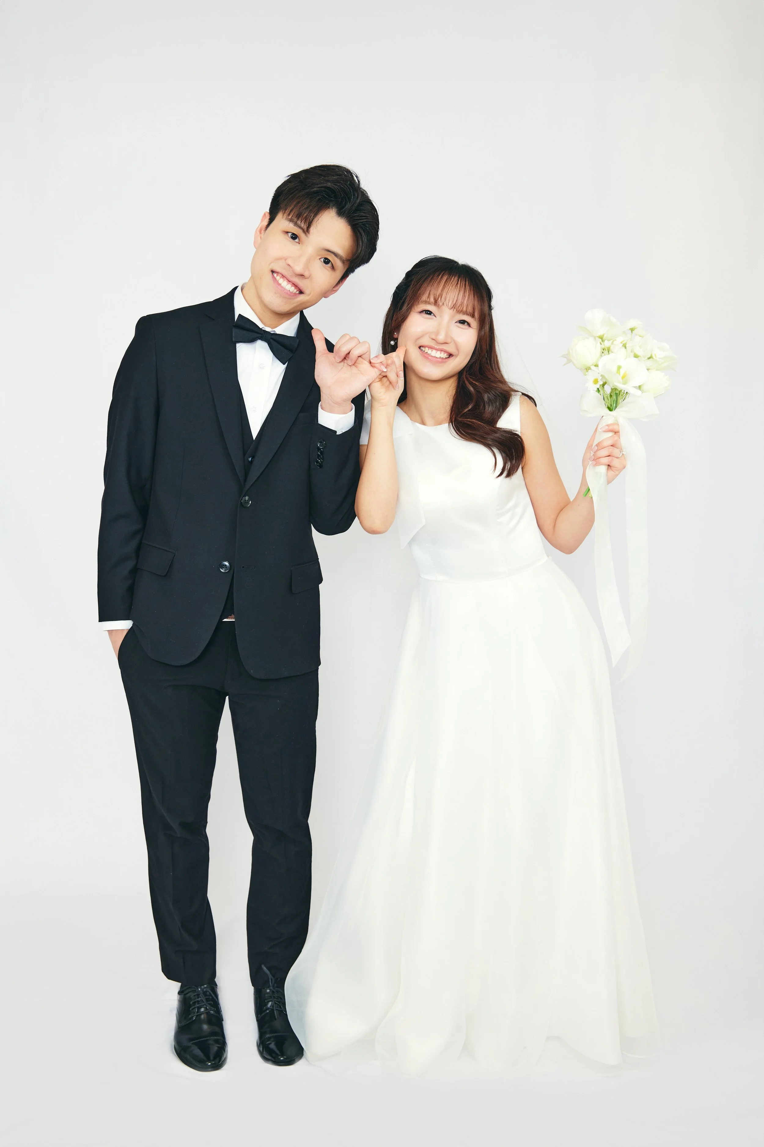 A happy couple in wedding attire posing for a photo against a white background, the groom dressed in a black tuxedo and the bride in a white wedding dress holding a bouquet of white flowers.
