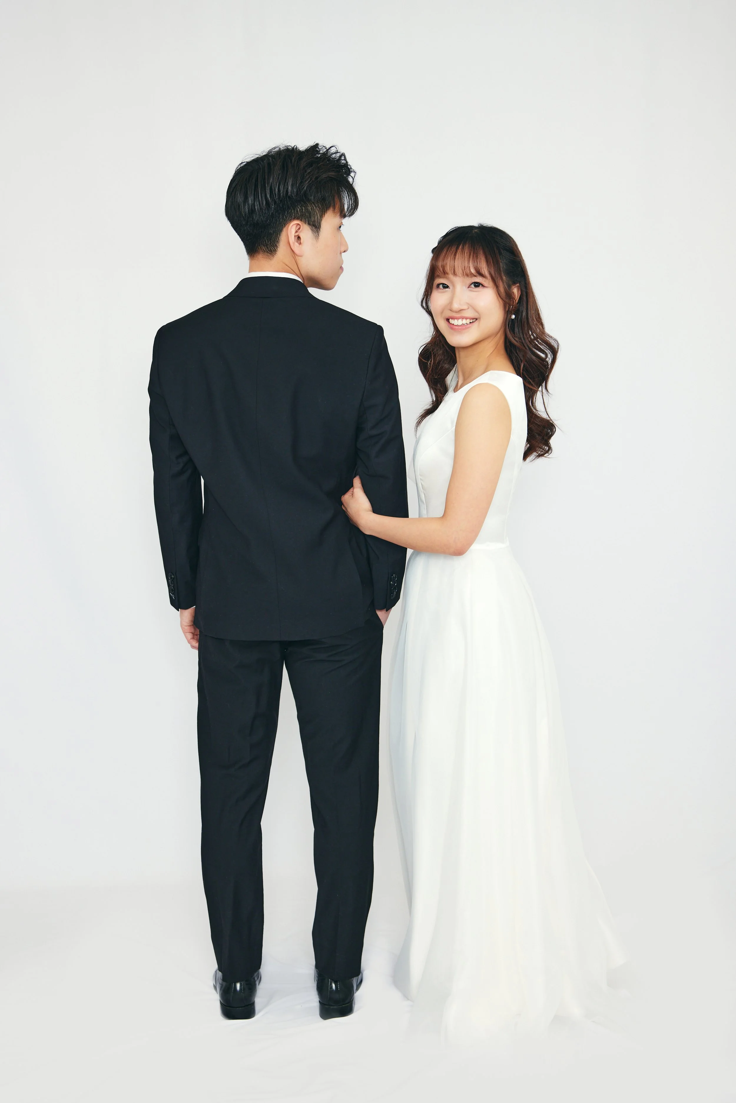 A smiling woman in a white dress holding onto a man in a black suit on a white background.