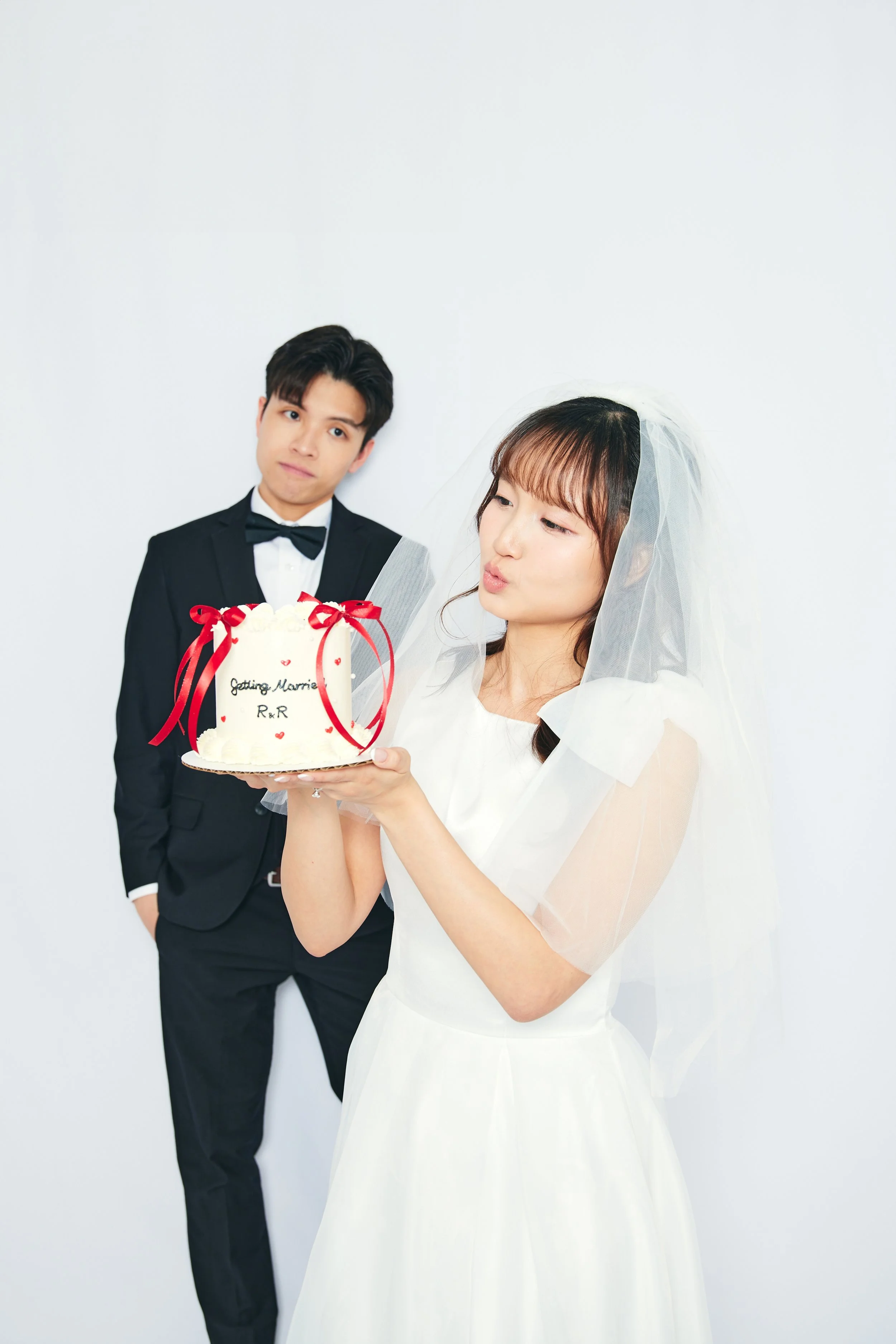 A bride in a white wedding dress and veil is holding a cake with red ribbons and hearts, reading 'Getting Married R.R'. A groom in a black tuxedo stands behind her, watching.