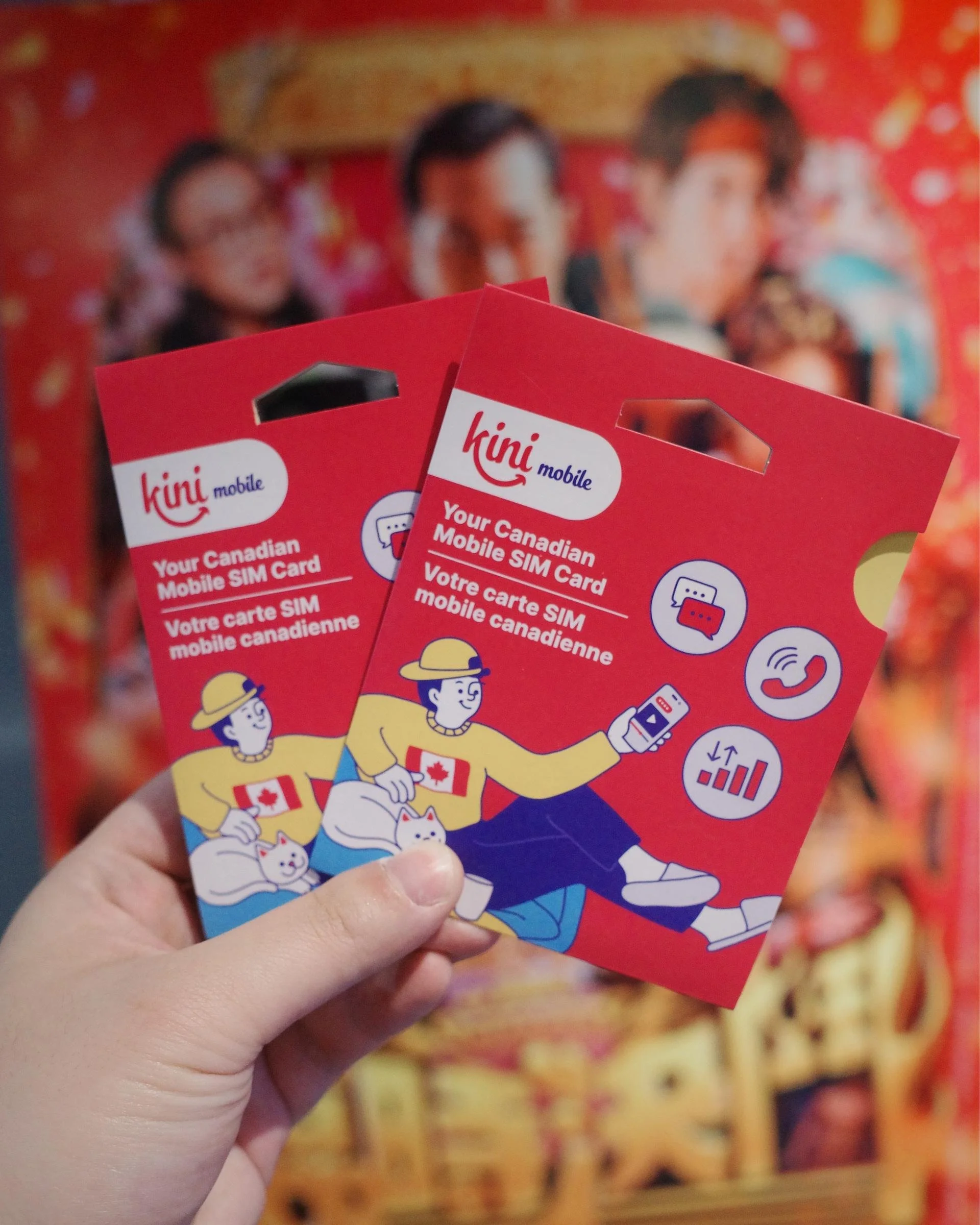 A hand holding two kini mobile SIM card packs in front of a film poster.