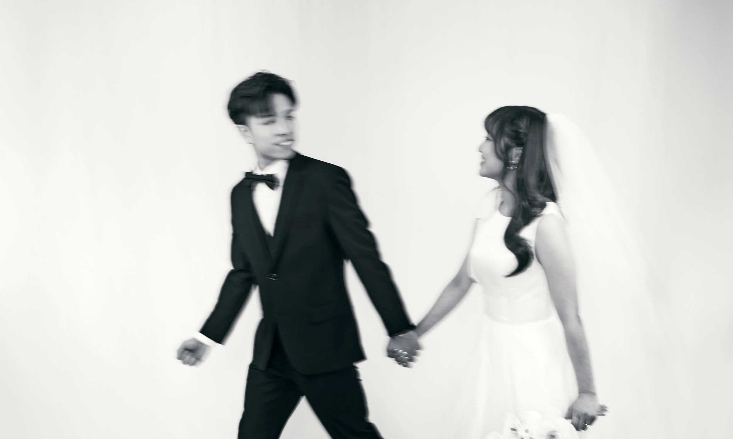 A black and white photo of a bride and groom holding hands, with the bride wearing a wedding dress and the groom in a tuxedo, walking together.