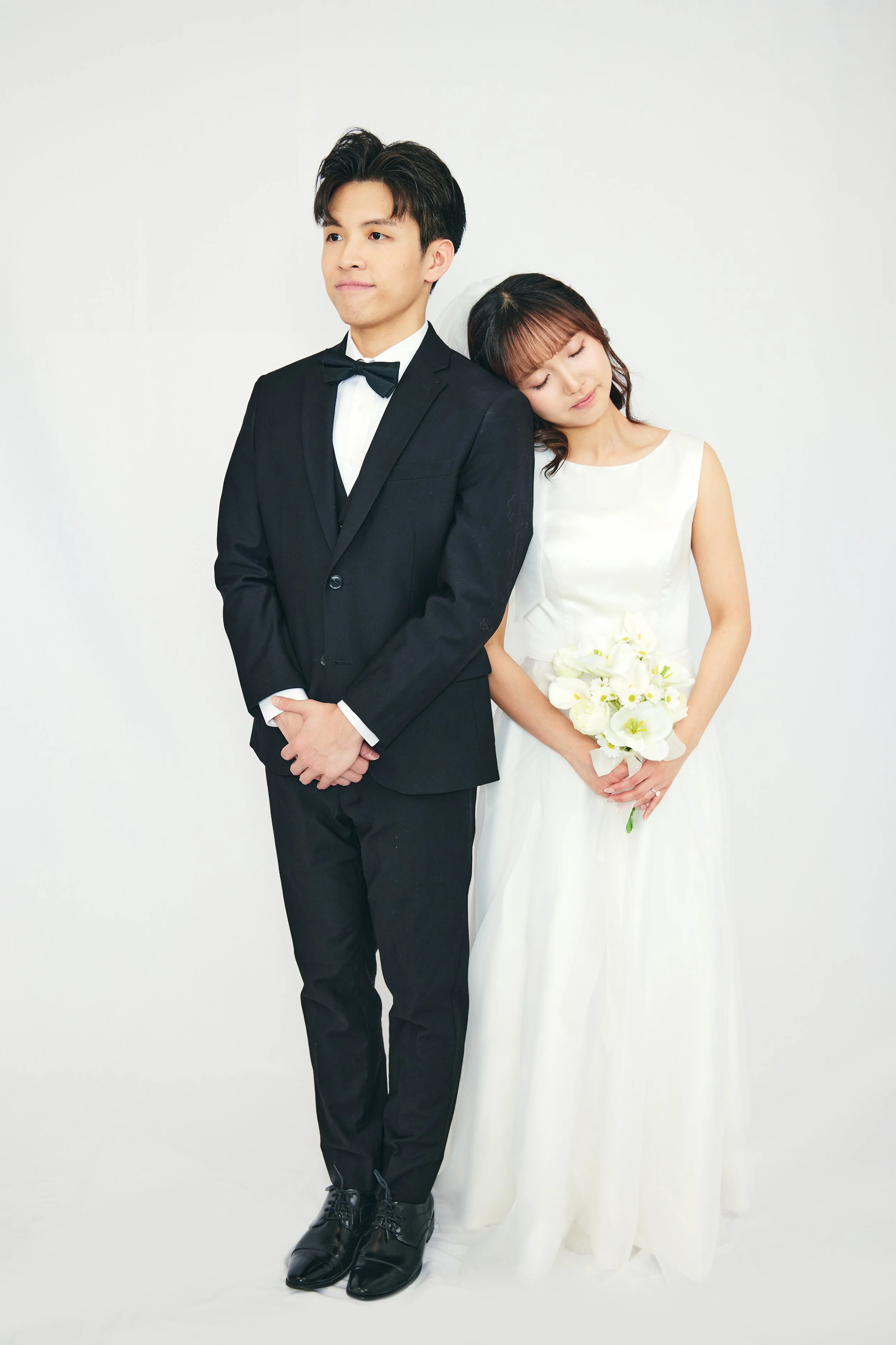 A bride and groom stand together against a white background; the groom is in a black tuxedo with a bow tie, and the bride is in a white wedding dress holding a bouquet of white flowers, with the bride resting her head on the groom's shoulder.