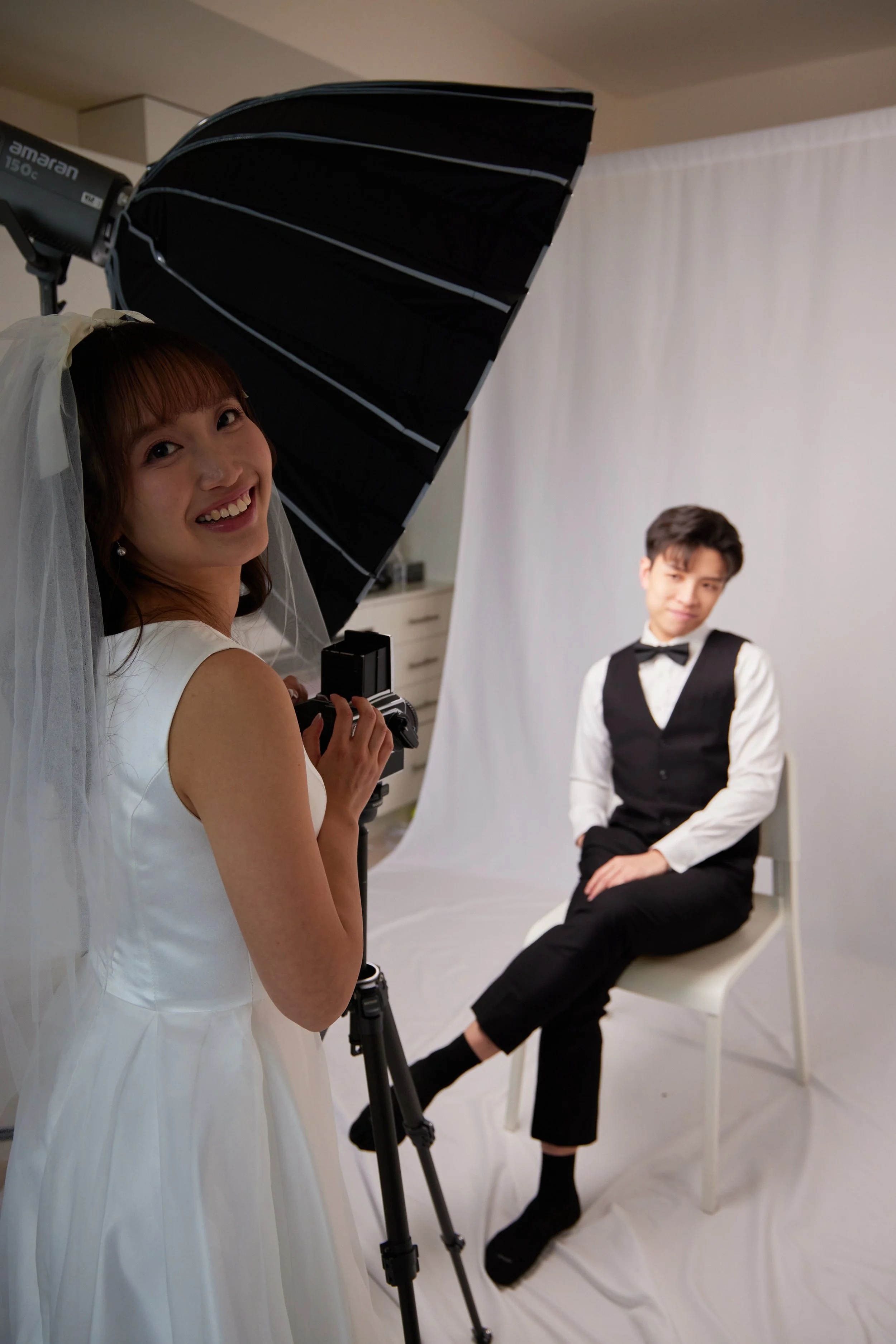 A woman in a white dress and veil is taking a photo of a man in a tuxedo sitting on a chair in a photo studio with a white backdrop.