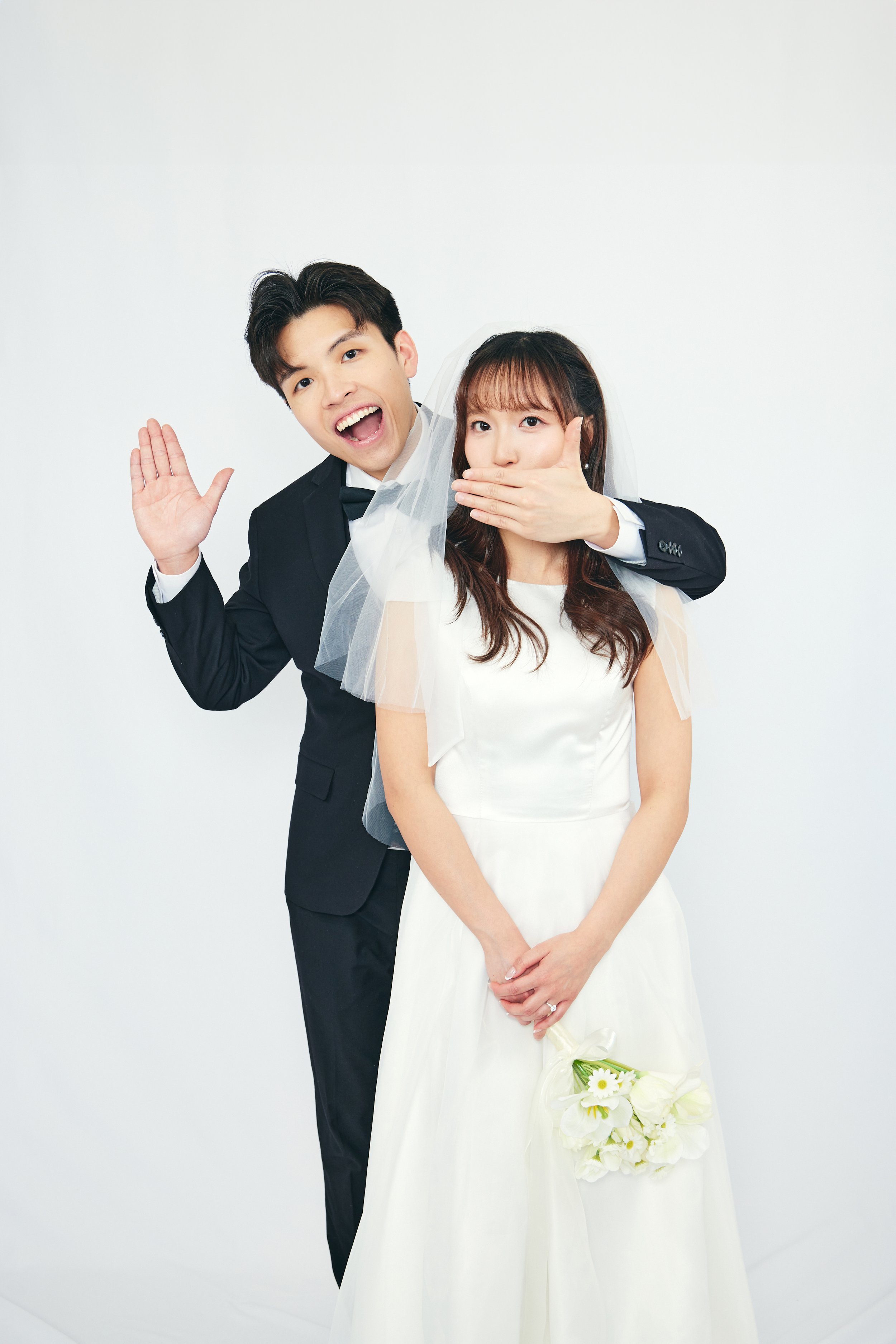 A bride and groom in wedding attire posing playfully against a plain white background. The groom is smiling and waving, while the bride has a surprised expression as the groom covers her mouth with his hand. The bride holds a small bouquet of white f