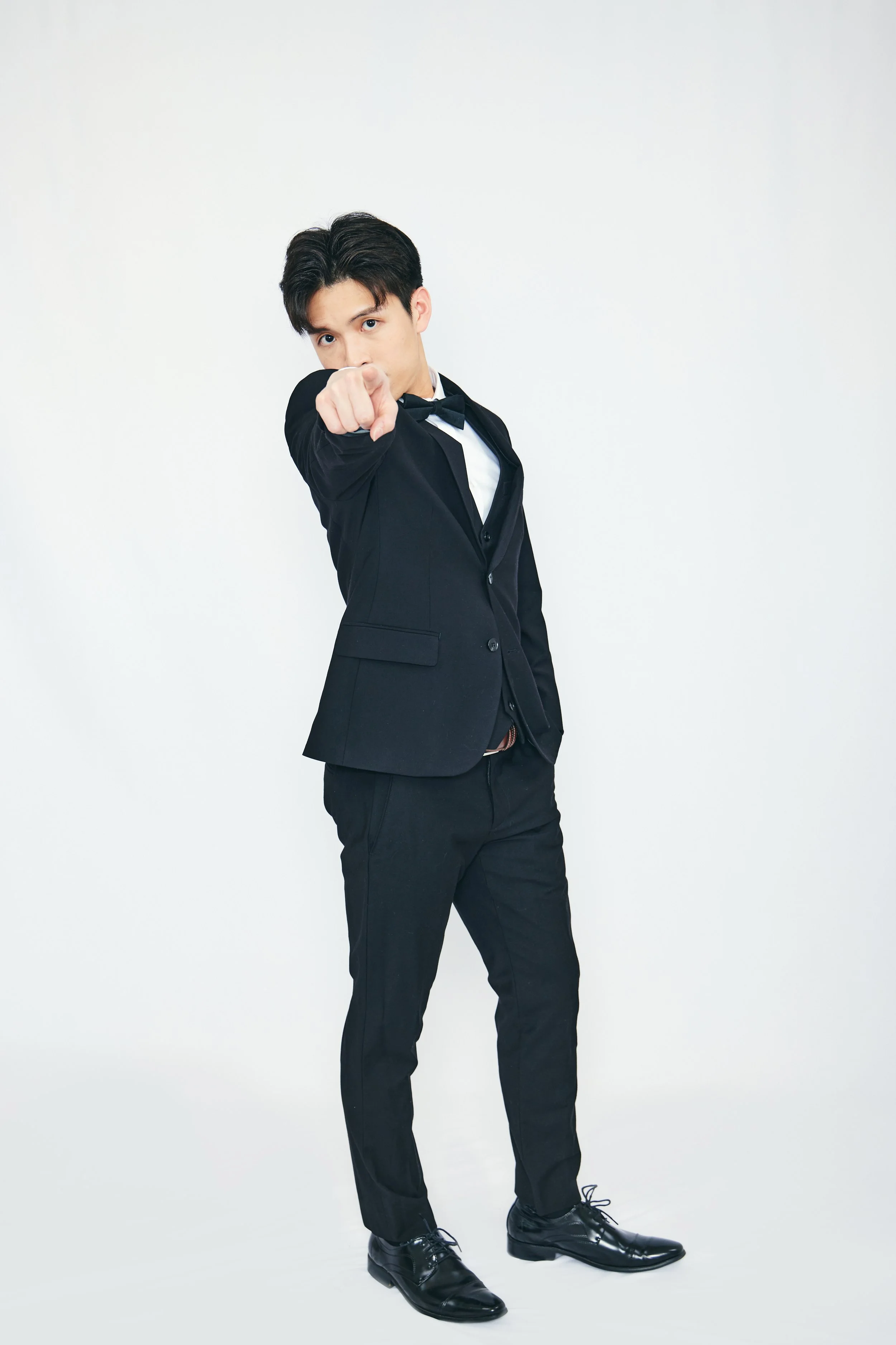 Young man in black tuxedo pointing at the camera on white background.