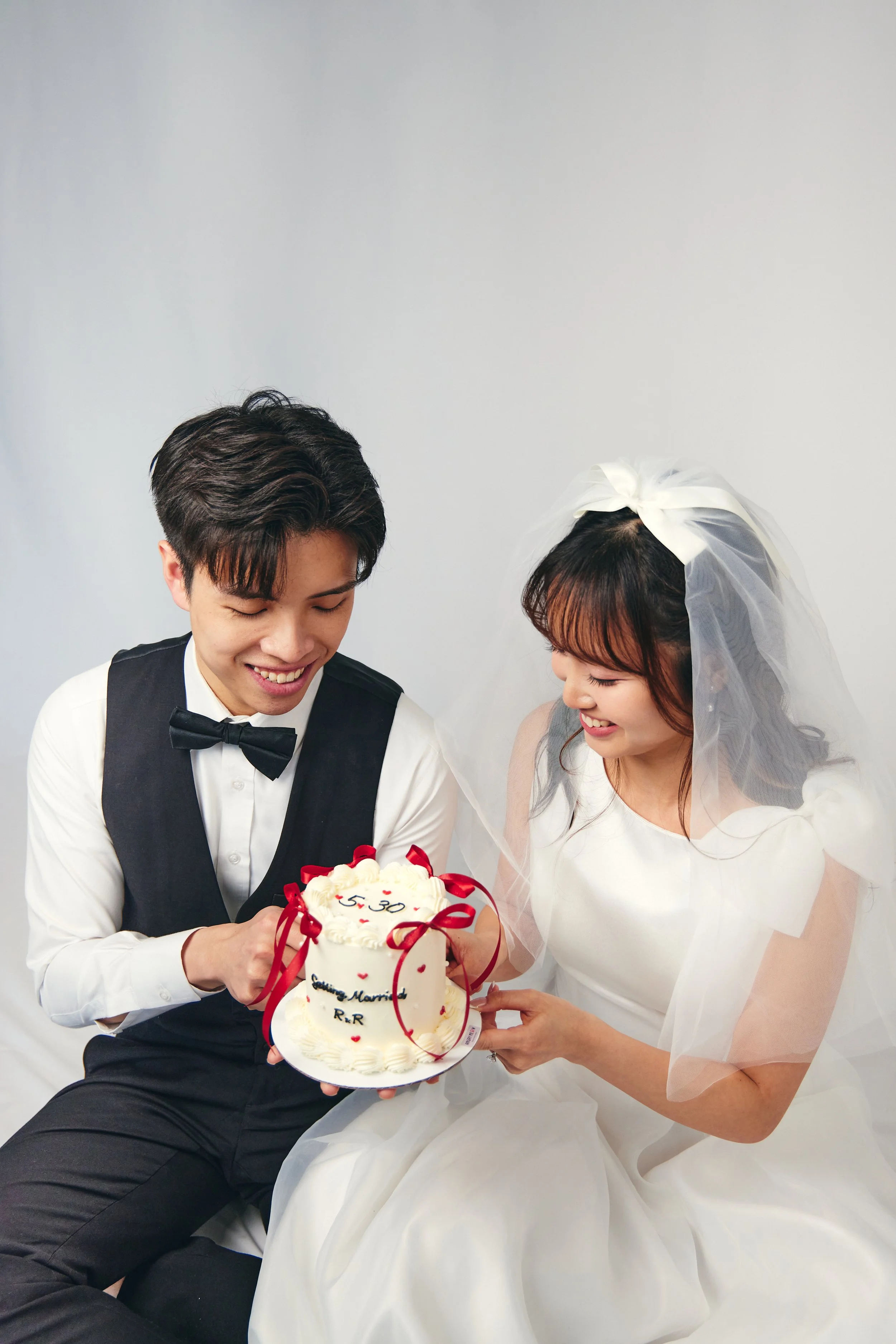A bride and groom in wedding attire holding a small wedding cake decorated with red ribbons and the date 5.30, smiling and looking at the cake.