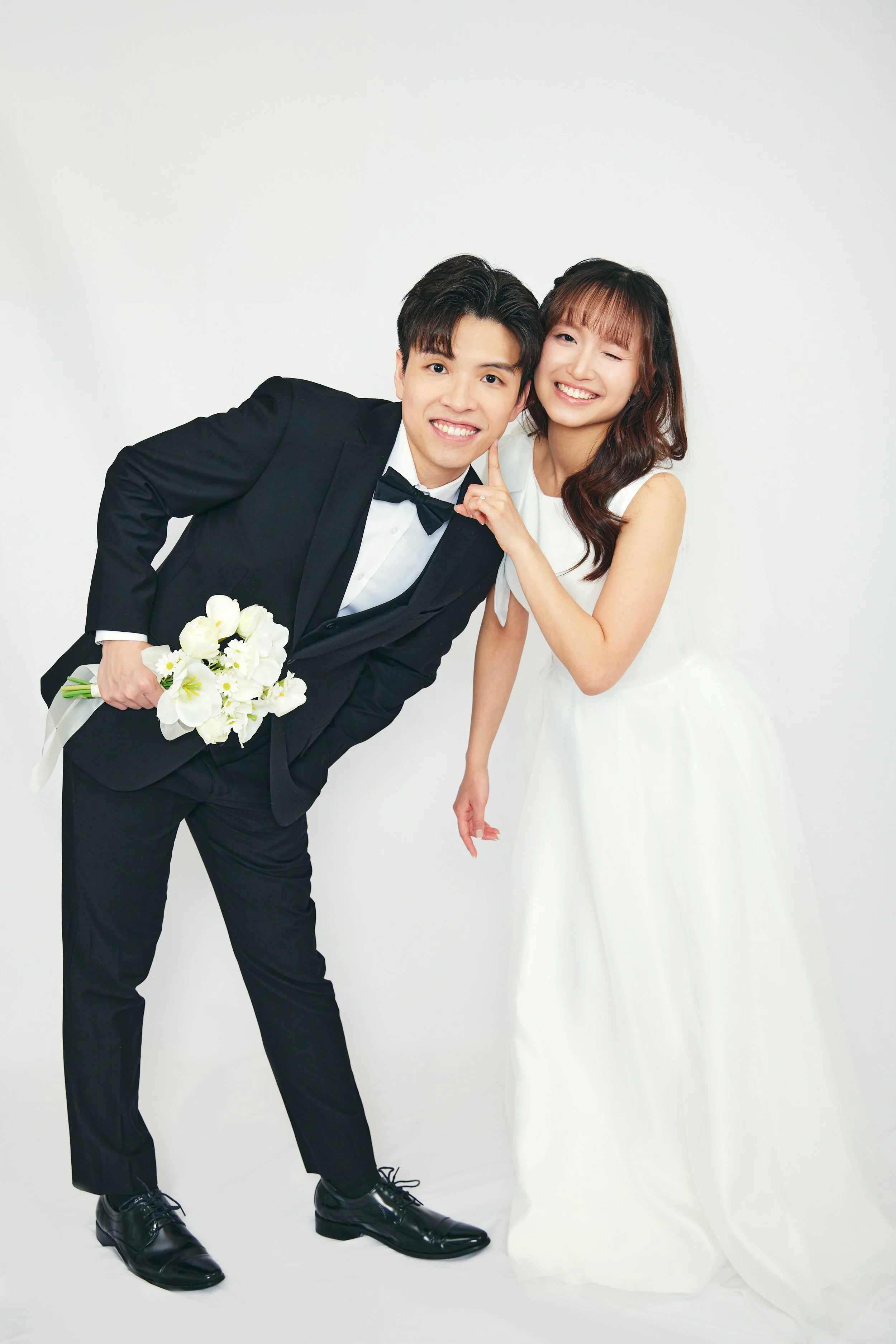 A couple dressed in wedding attire, with the groom in a black tuxedo and the bride in a white wedding dress, smiling and posing in front of a plain white background.