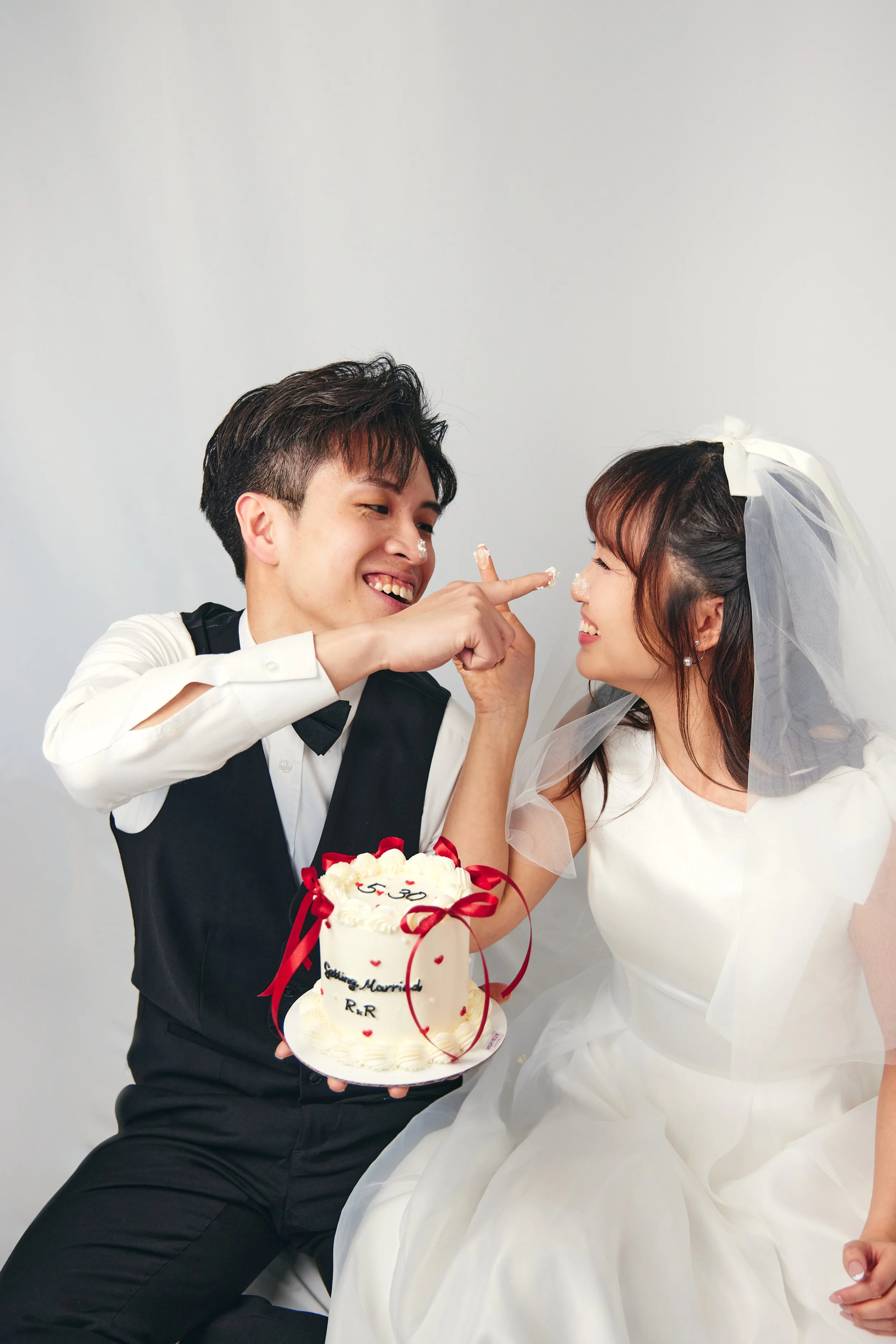 A happy bride and groom celebrating their wedding, with the groom playfully touching the bride's face. The groom is holding a wedding cake decorated with red ribbons and romantic inscriptions. The bride is wearing a white wedding dress and a bridal v