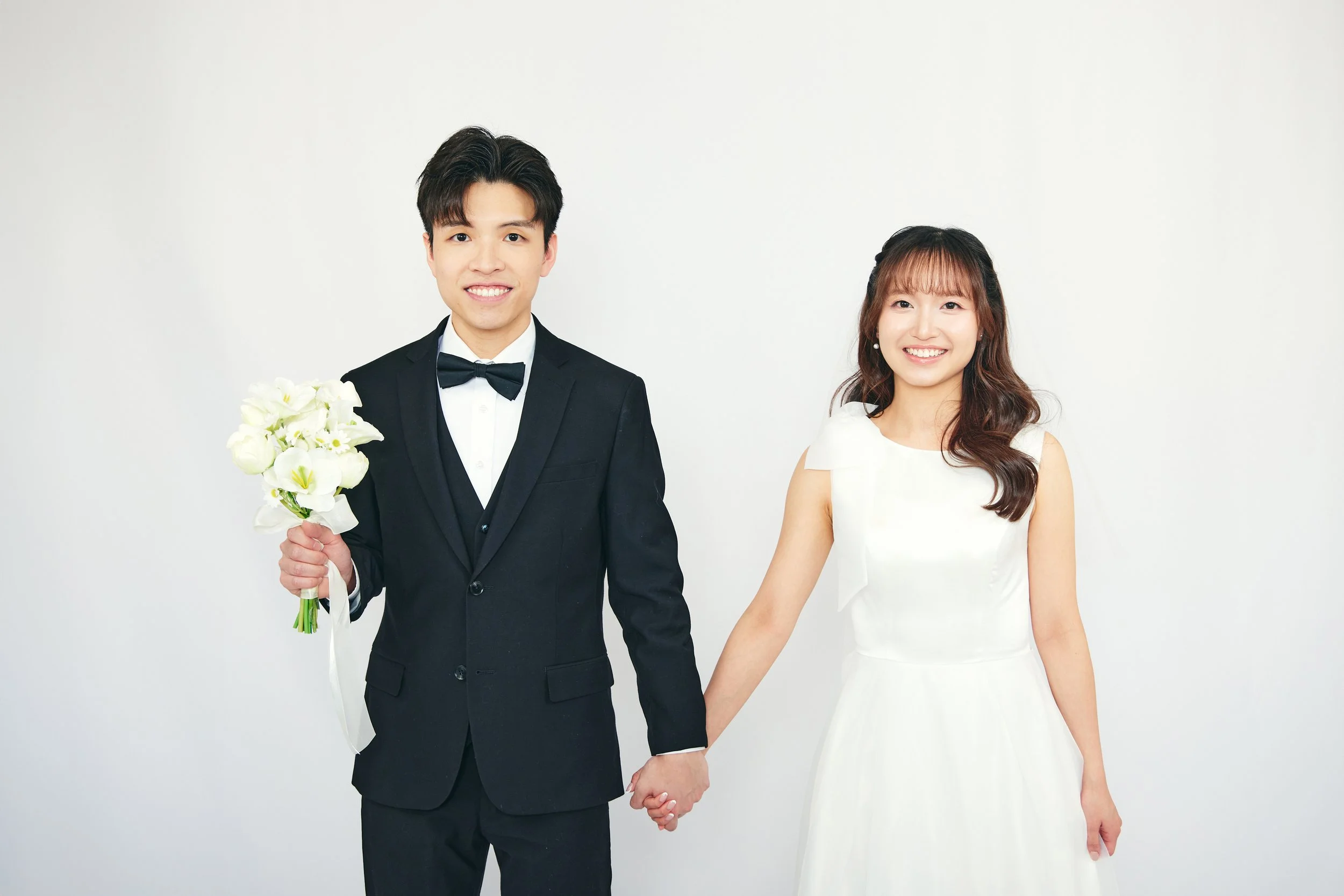 A newlywed couple holding hands, with the groom holding a bouquet of white flowers, standing against a plain white background.