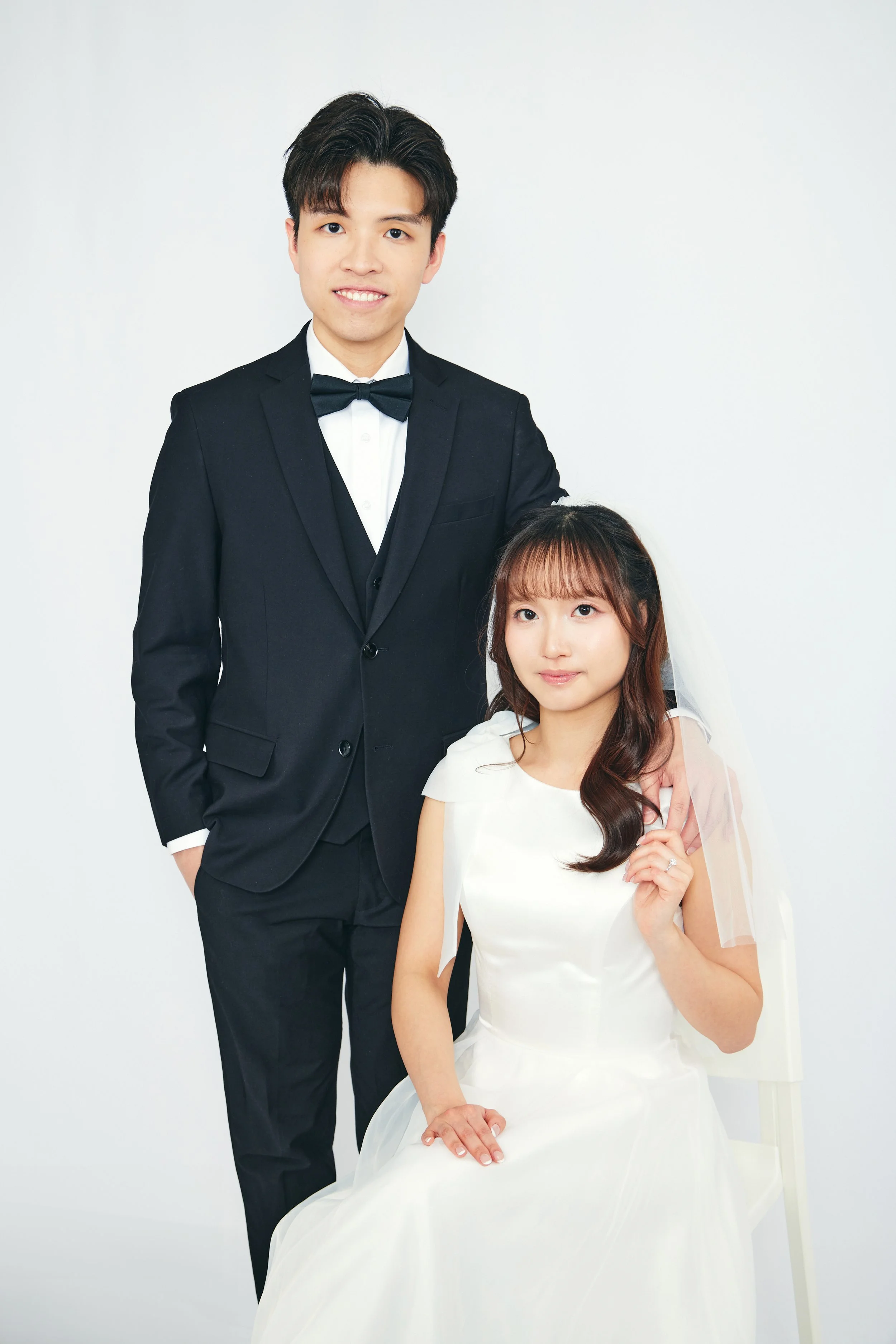 A man in a tuxedo standing next to a woman in a wedding dress with a veil. They are posing against a plain white background.