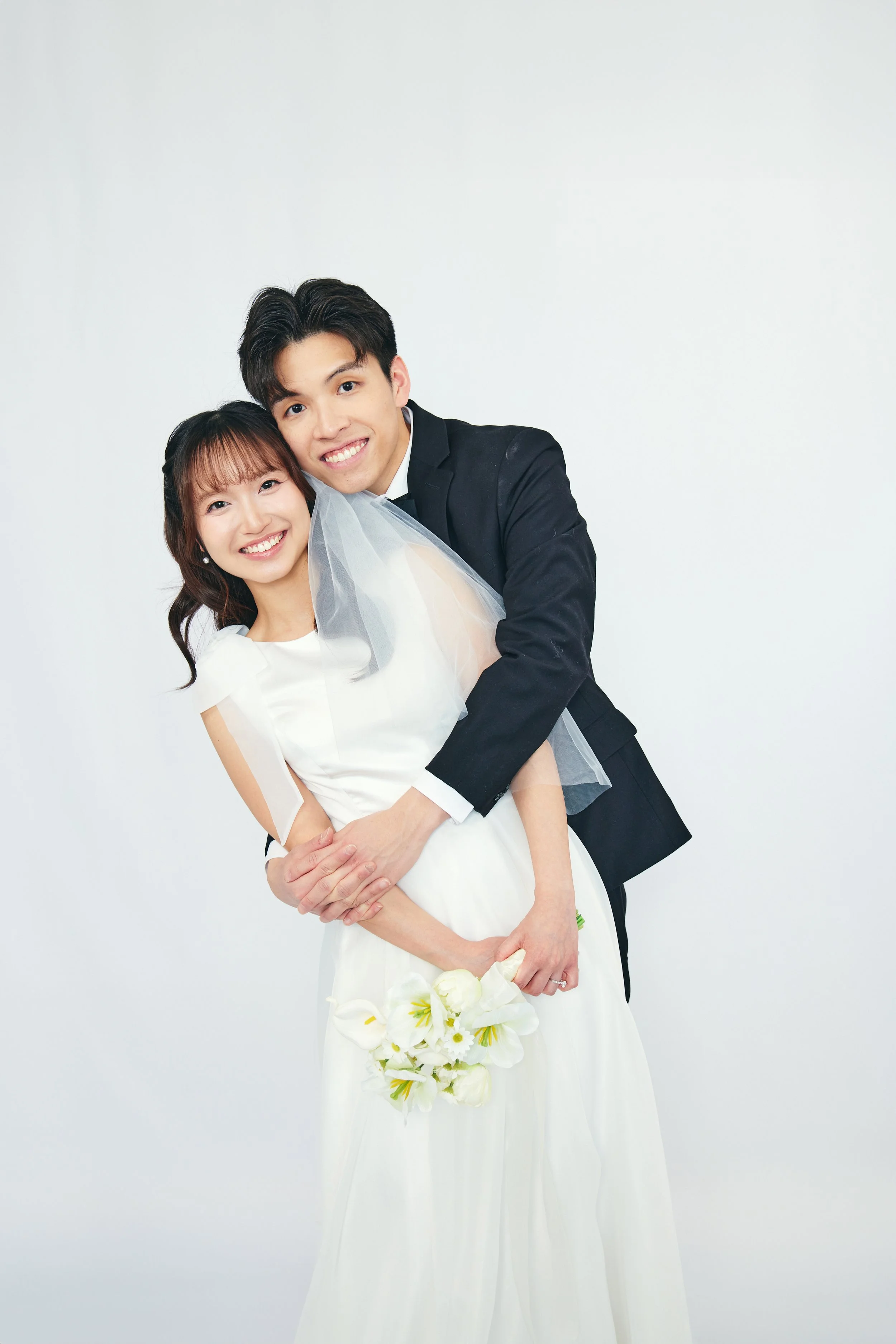 A happy couple dressed in wedding attire posing together against a plain white background, with the woman holding a white floral bouquet.