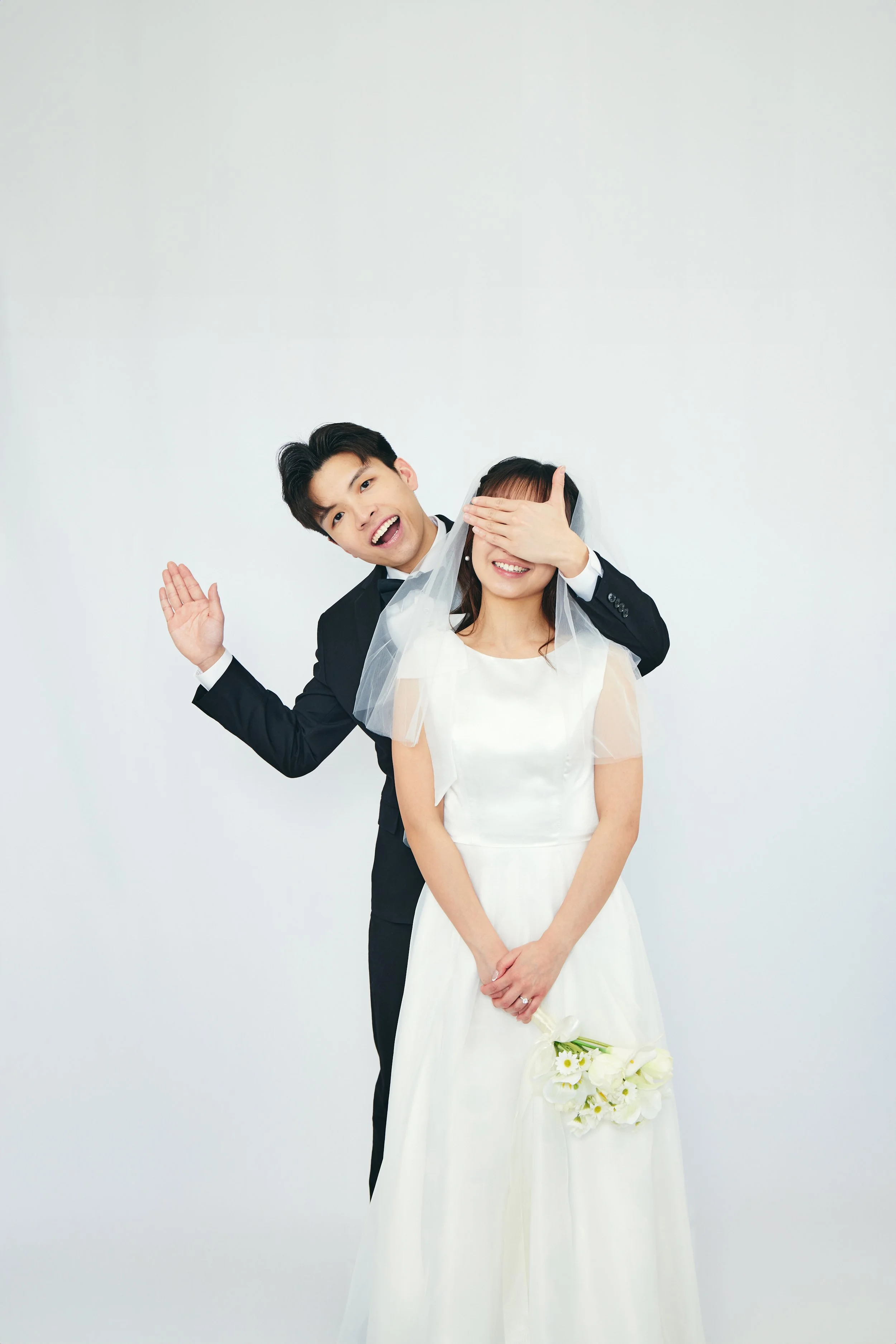 A smiling bride holding a bouquet of white flowers and a groom covering her eyes with his hand during a wedding photo shoot.