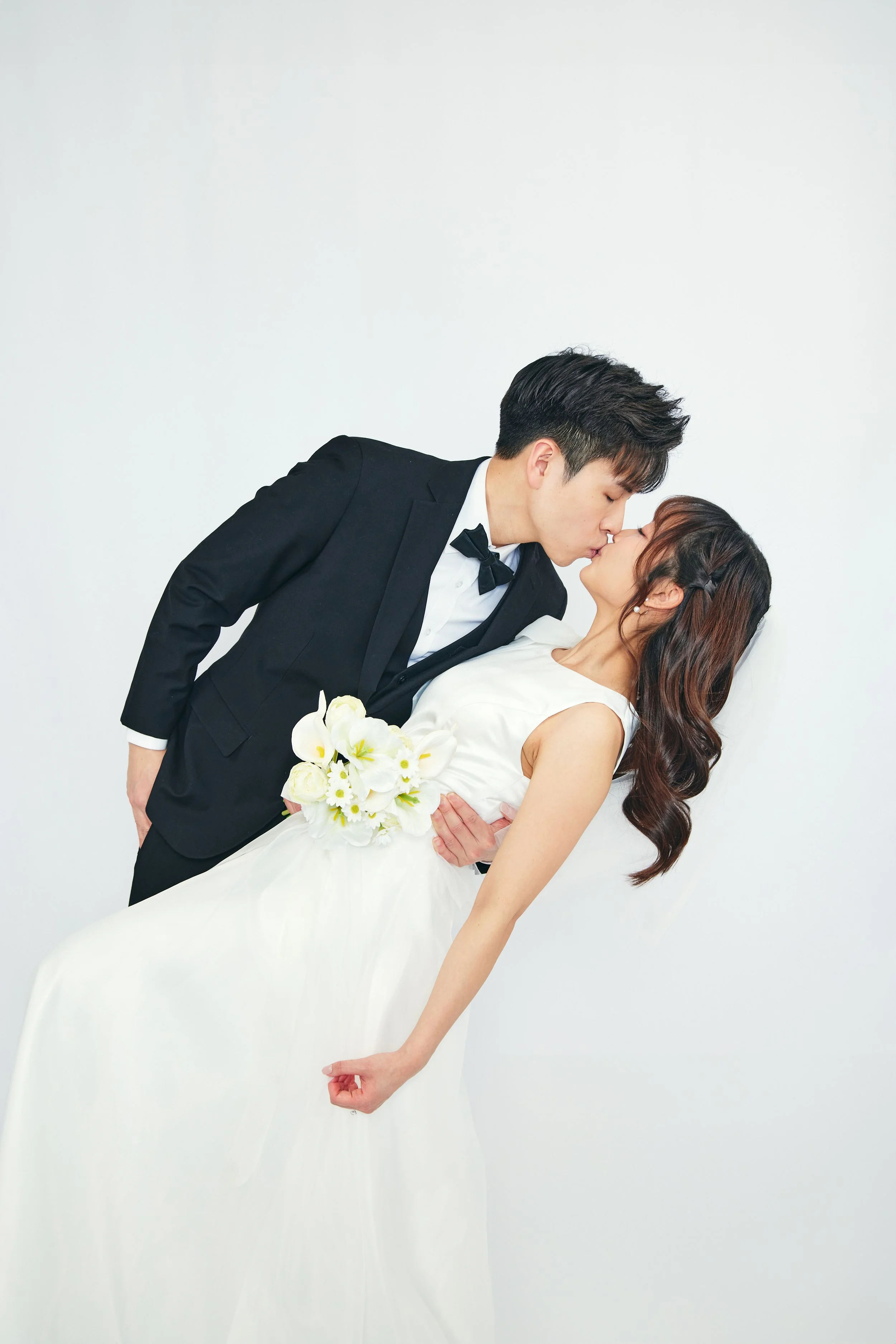A couple in wedding attire sharing a kiss, with the groom in a black tuxedo and the bride in a white wedding dress holding a bouquet of white flowers.
