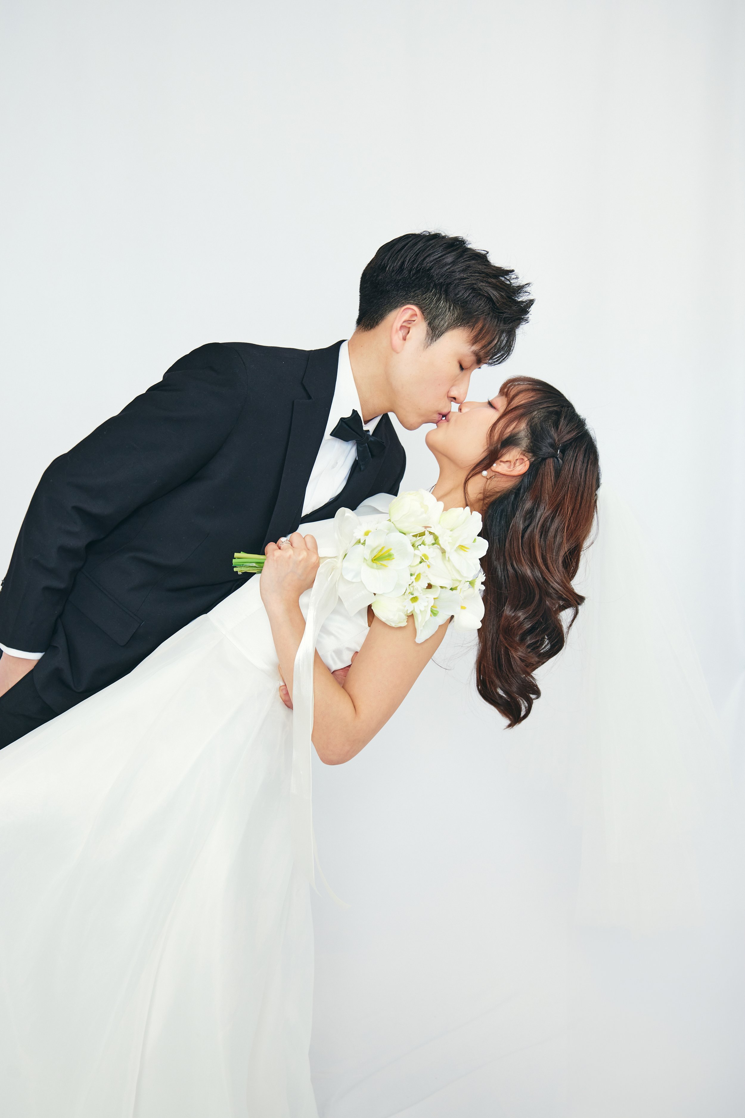 A bride and groom kiss, with the groom in a black tuxedo and bow tie, and the bride in a white wedding gown holding a bouquet of white flowers, against a plain white background.