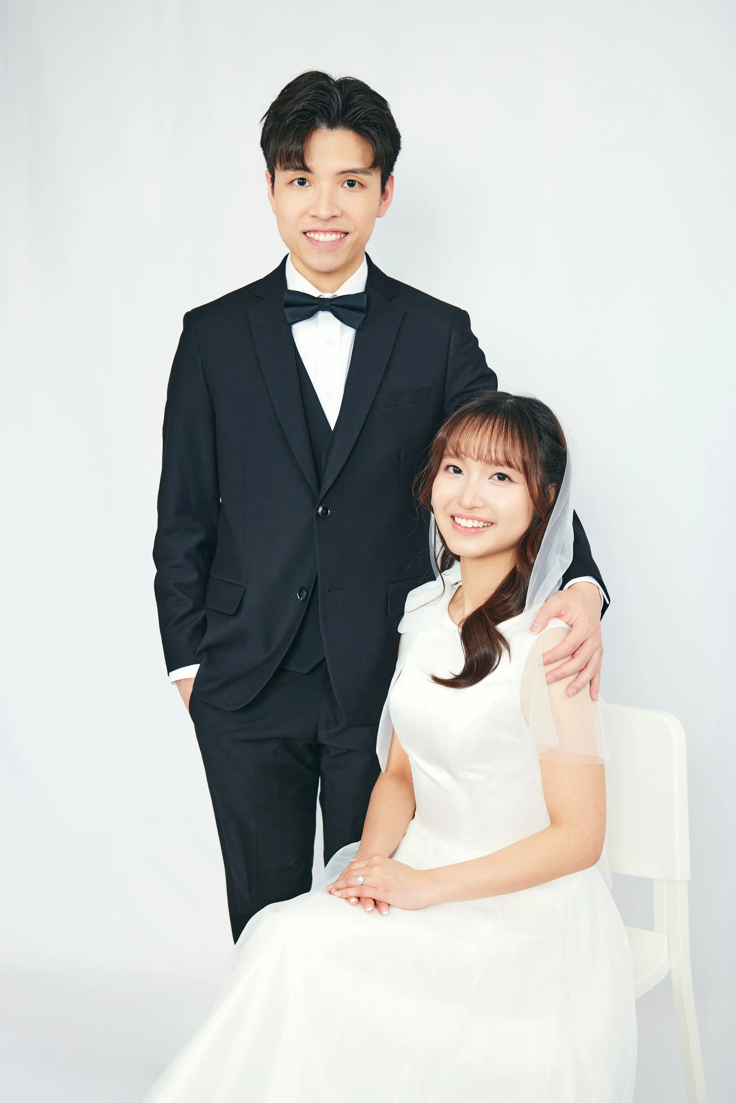 A newlywed couple dressed in wedding attire posing against a white background, with the groom standing and the bride seated, both smiling.