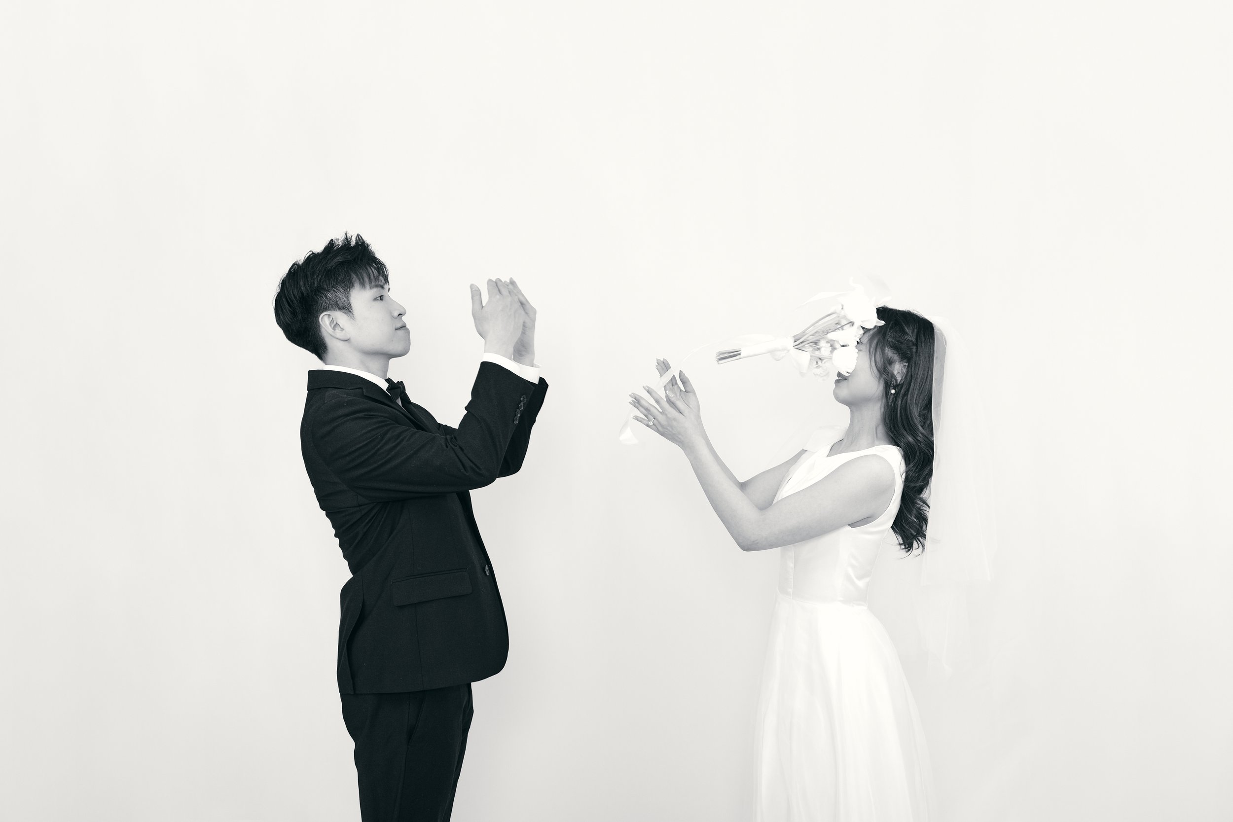 A black and white photo of a wedding scene with a man in a suit clapping while a woman in a wedding dress throws a bouquet of flowers into the air.