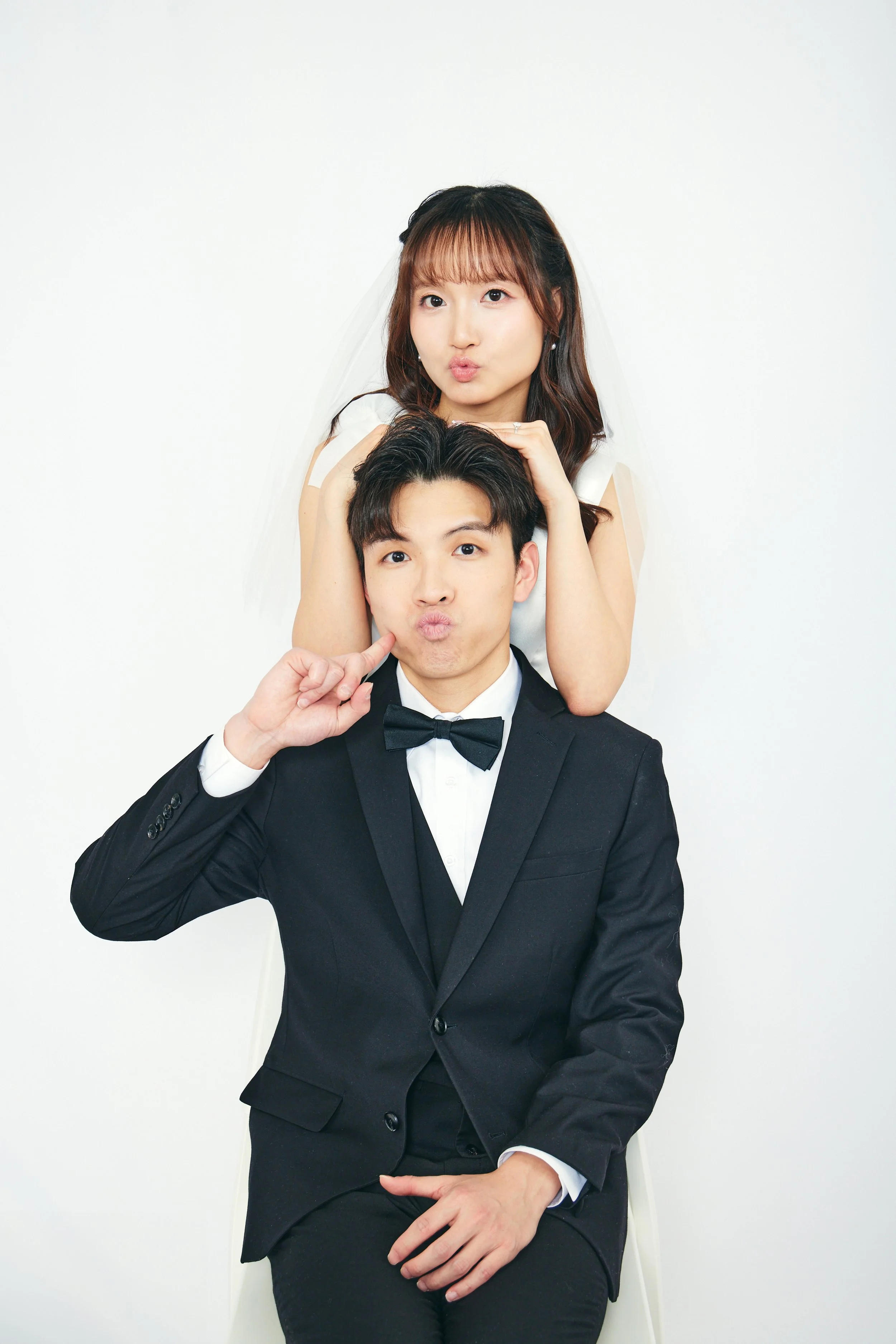 A couple in wedding attire making playful facial expressions, with the woman standing behind the man and resting her arms on his head, against a plain white background.