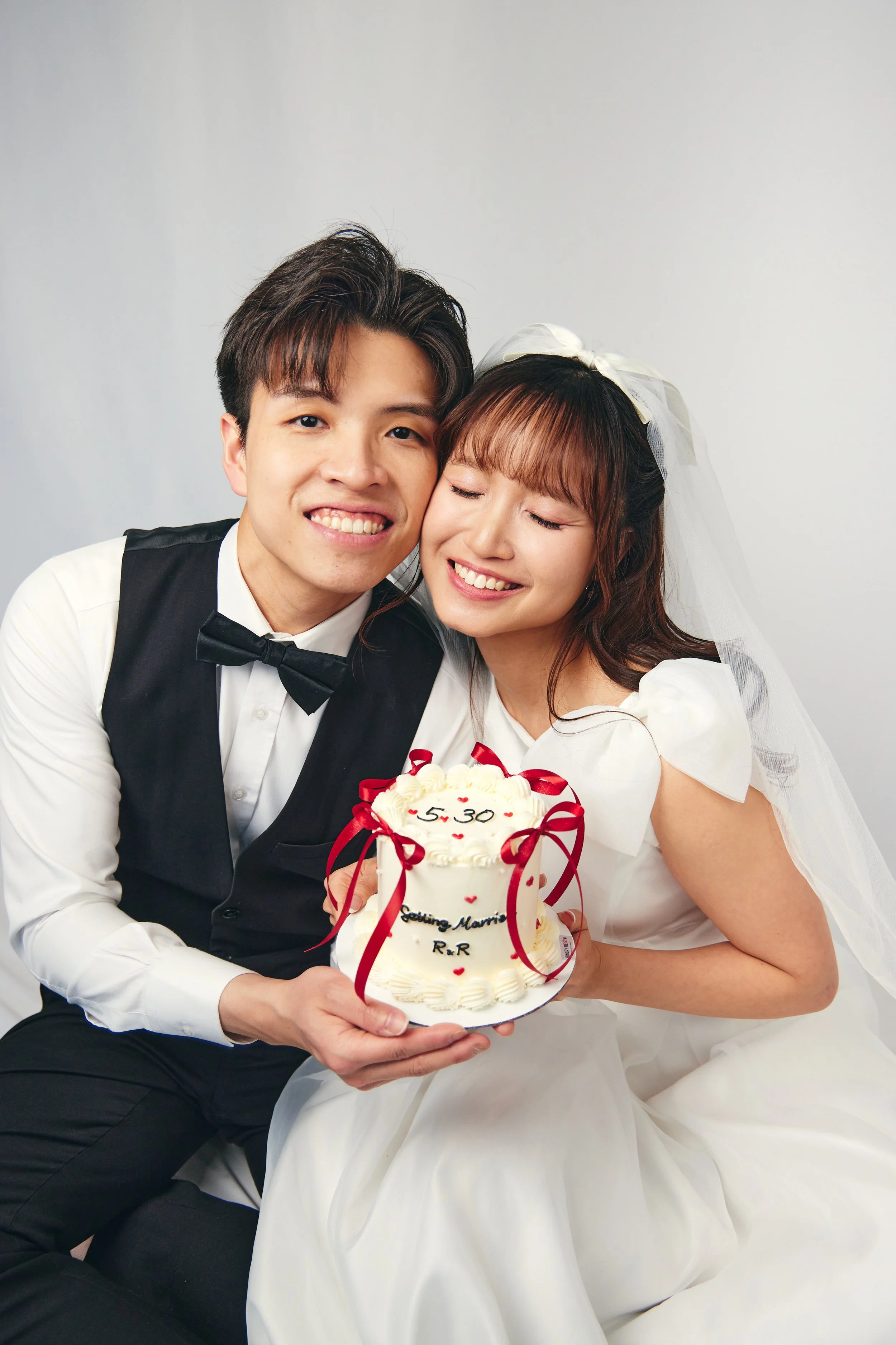A happy couple in wedding attire holding a decorated wedding cake, smiling with eyes closed, against a plain gray background.