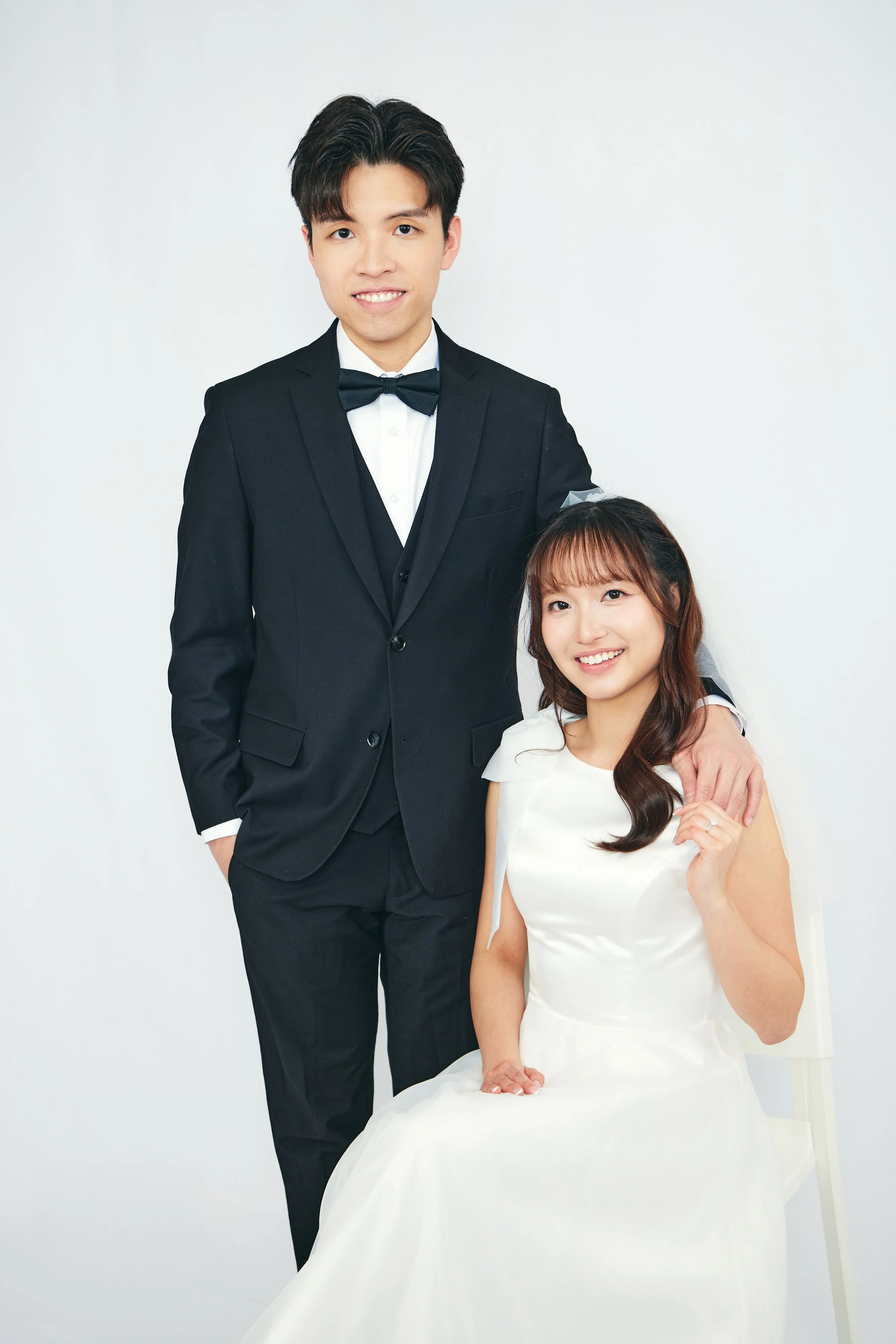 A happy couple at their wedding, with the groom in a black tuxedo and the bride in a white wedding dress, seated and standing against a plain white background.