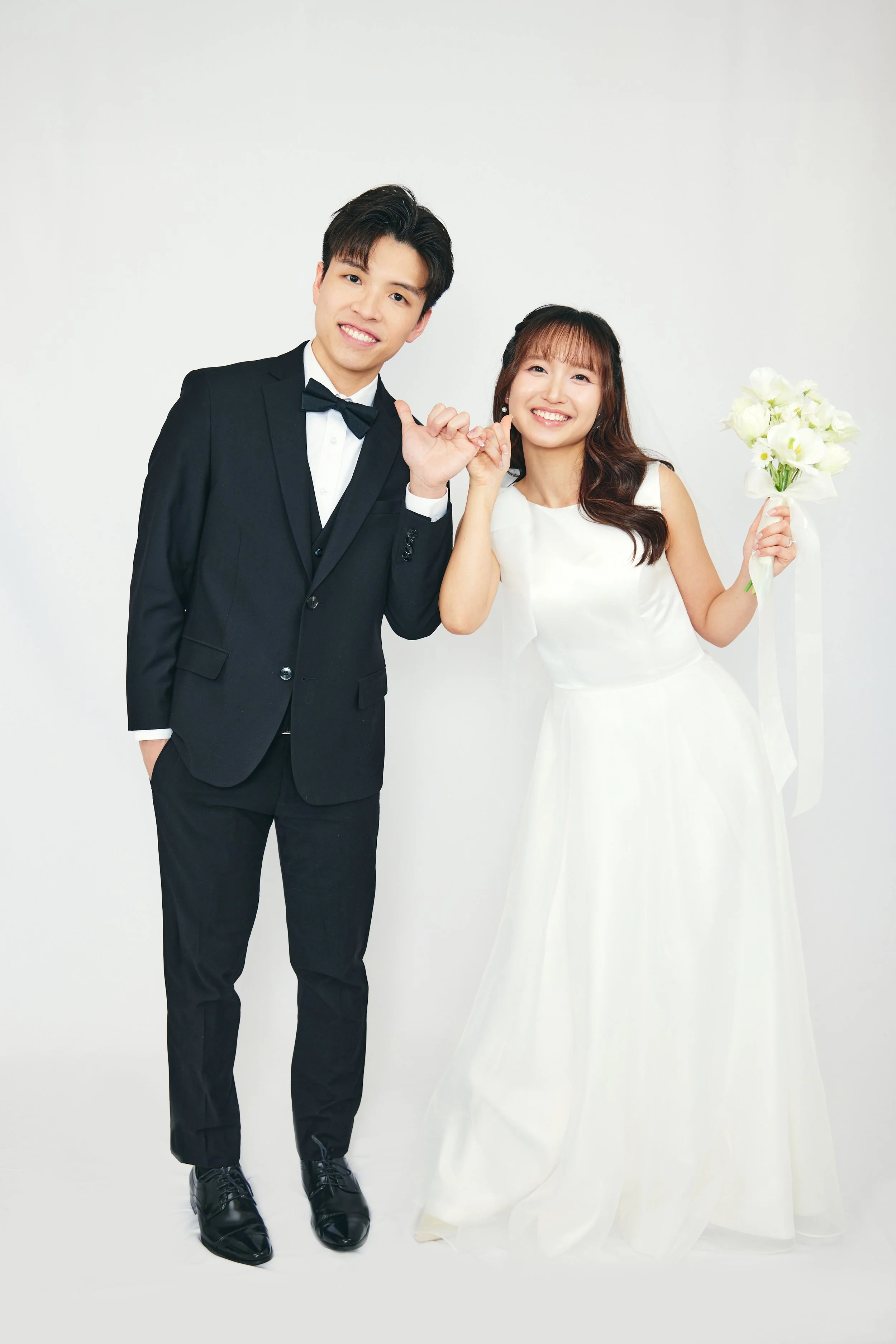 A newlywed couple dressed in wedding attire posing and smiling. The groom is in a black tuxedo with a bow tie, and the bride is in a white wedding gown holding a bouquet of flowers.