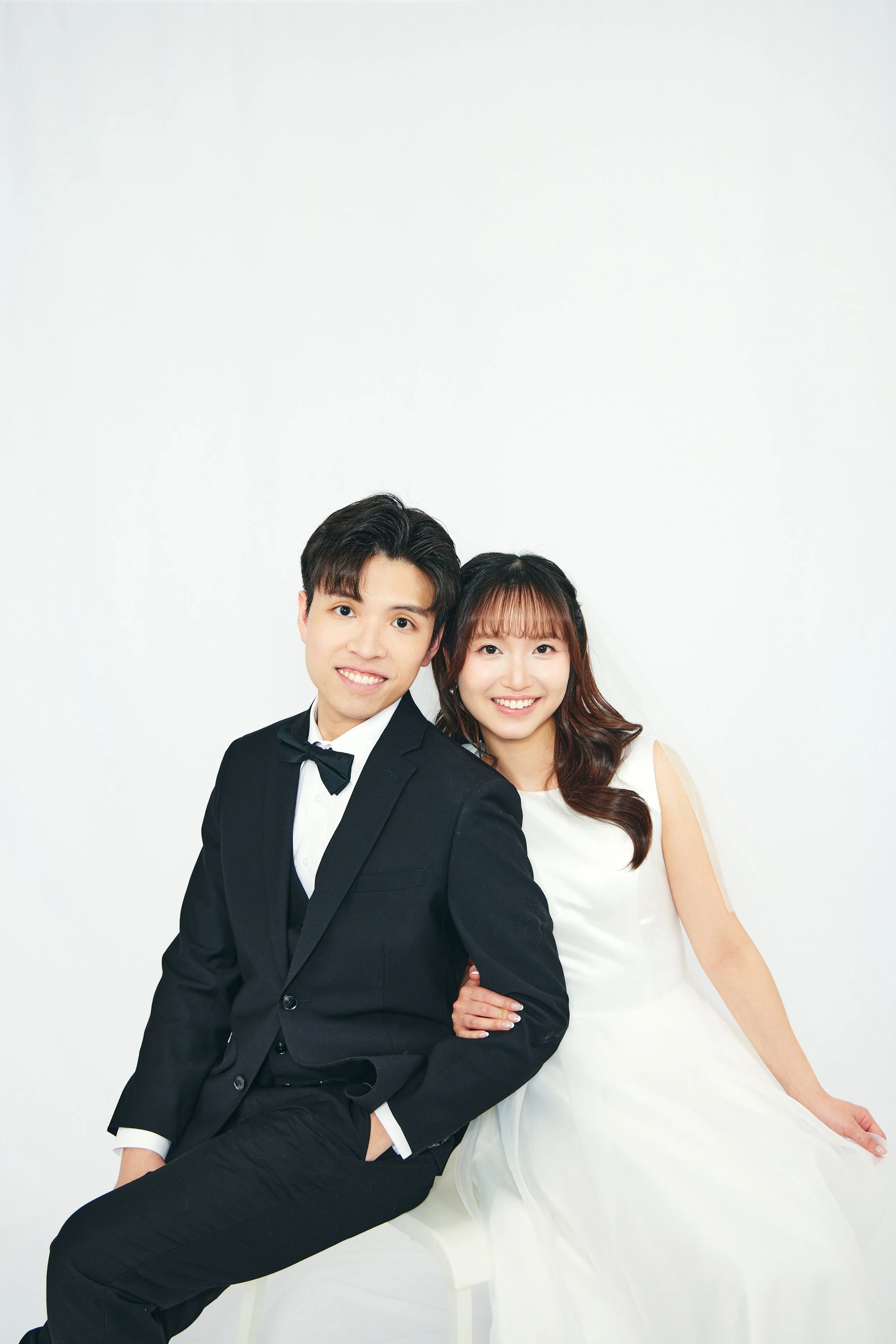 A young couple dressed in formal wedding attire, the man in a black tuxedo, and the woman in a white wedding dress, sitting together and smiling at the camera in front of a plain white background.