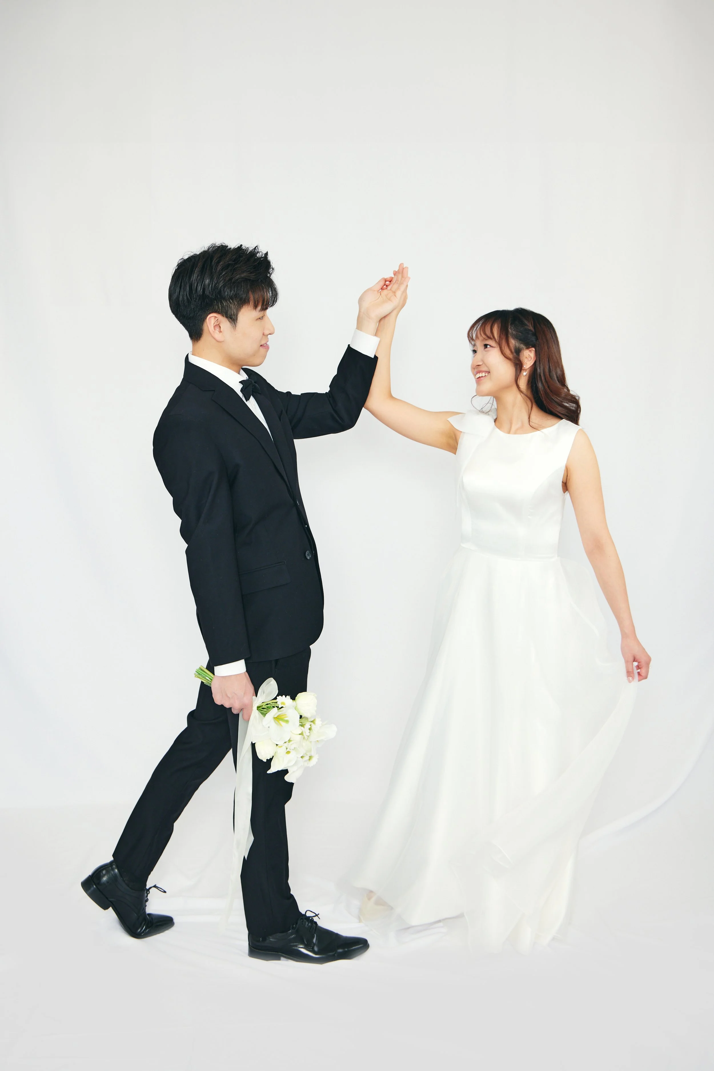 A bride and groom in wedding attire sharing a joyful moment, with the groom holding a bouquet of white flowers against a plain white background.