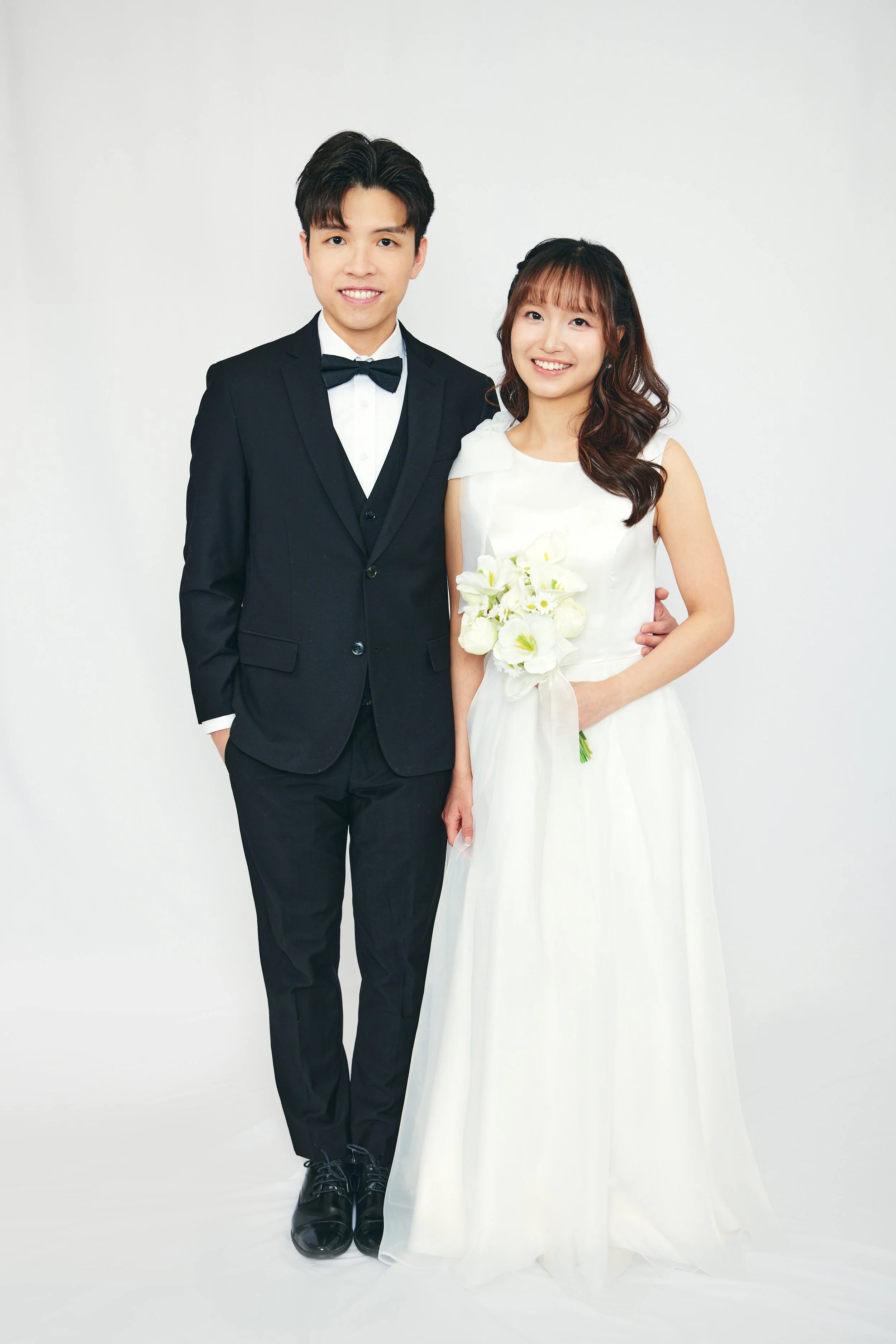 A young couple dressed in wedding attire, standing against a plain white background, smiling at the camera. The groom is in a black tuxedo with a bow tie, and the bride is in a white wedding dress holding a bouquet of white flowers.