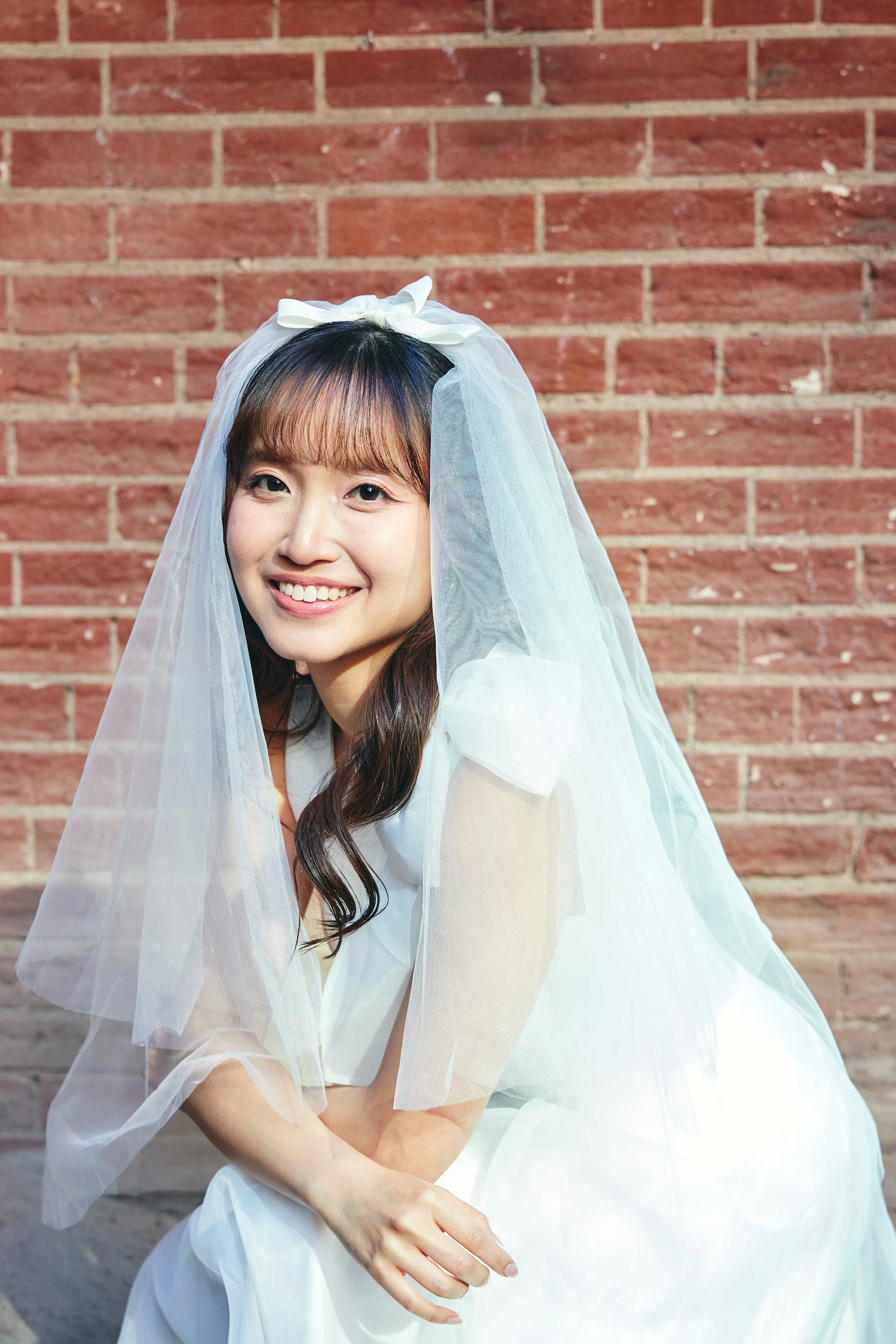 A young woman dressed as a bride, wearing a white dress and veil, smiling in front of a brick wall.