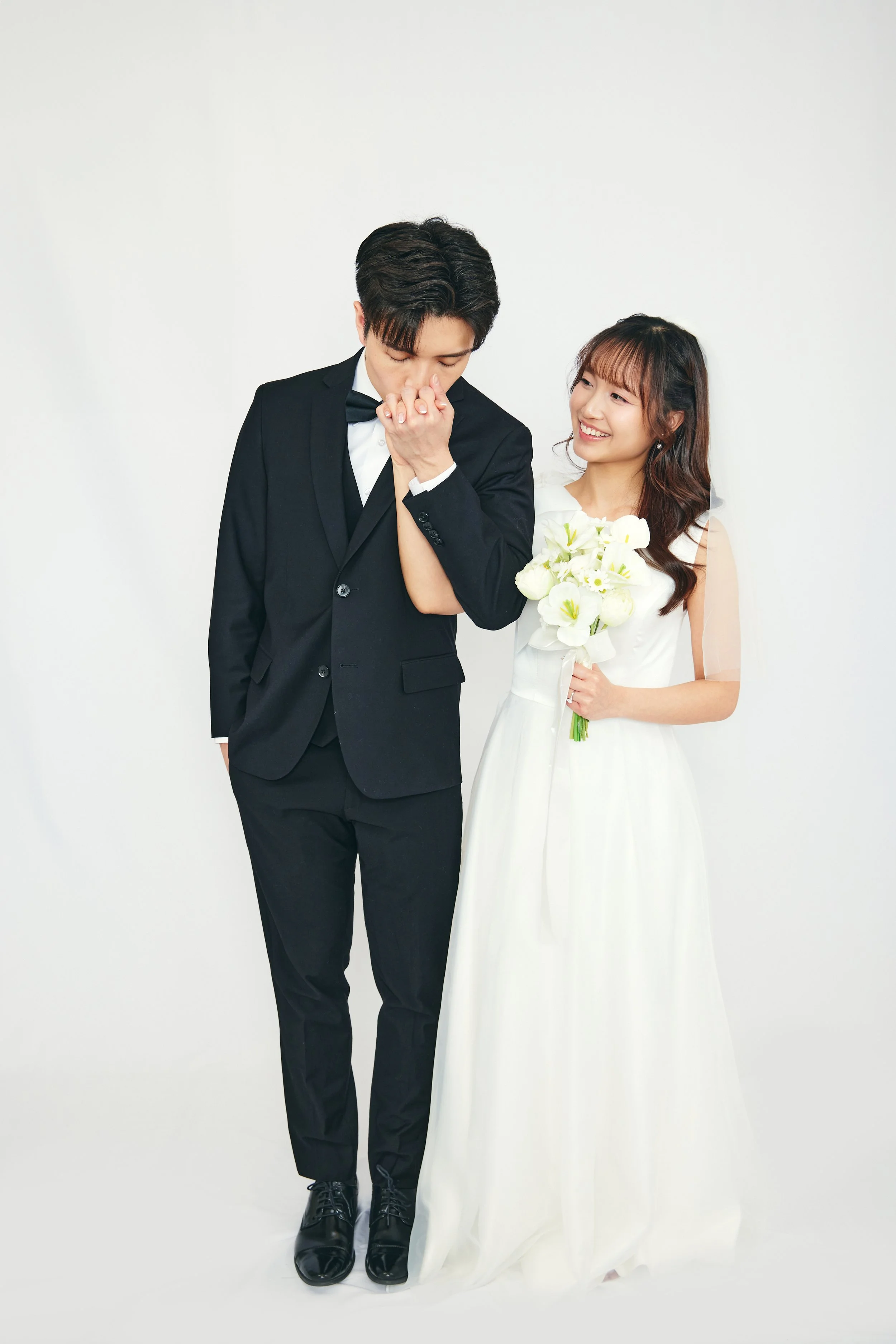 A bride in a white wedding dress holding a bouquet of white flowers standing next to a groom in a black tuxedo, both smiling in front of a plain white background.