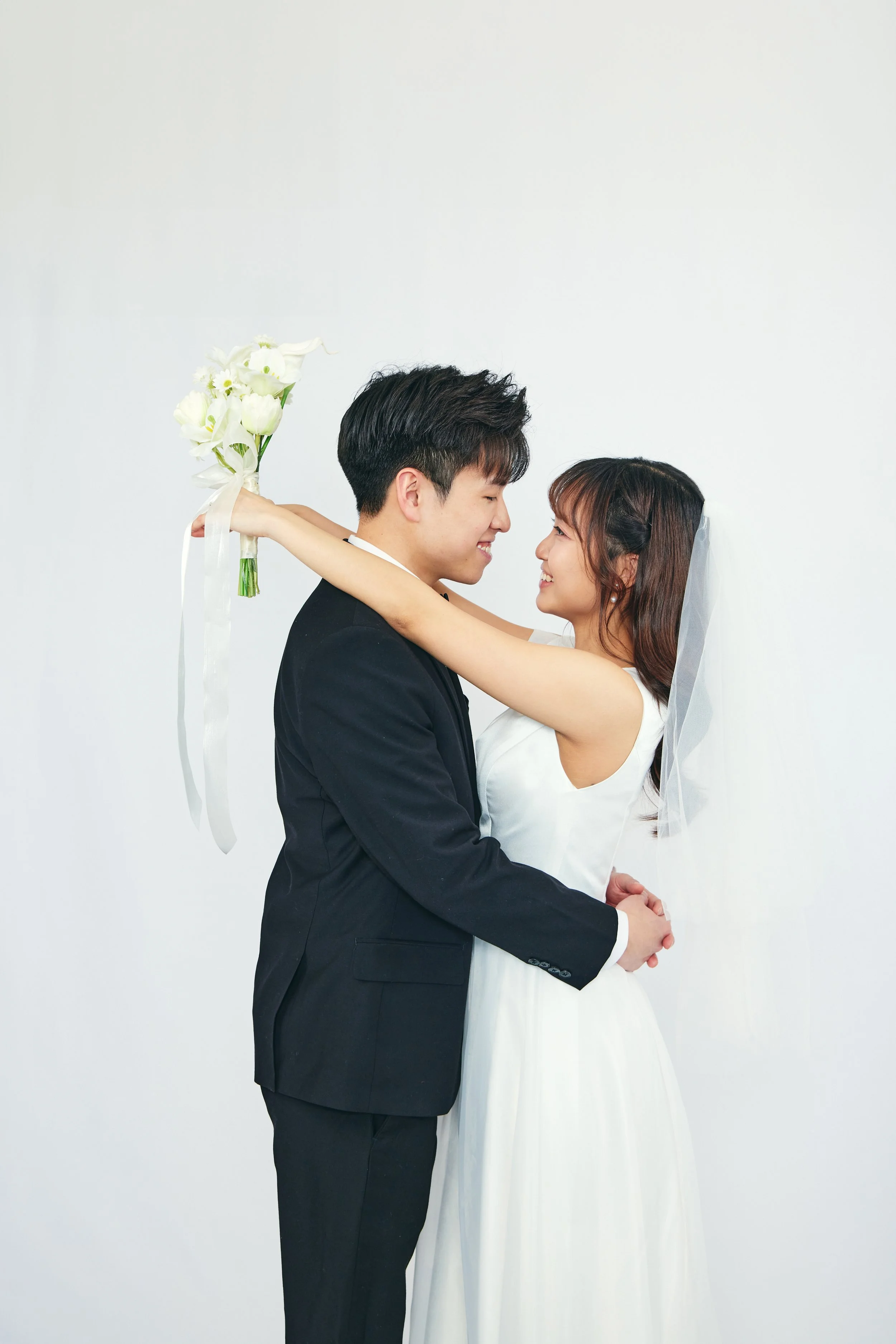 A happy bride and groom hold each other closely during their wedding photo shoot, with the bride holding a bouquet of white flowers.