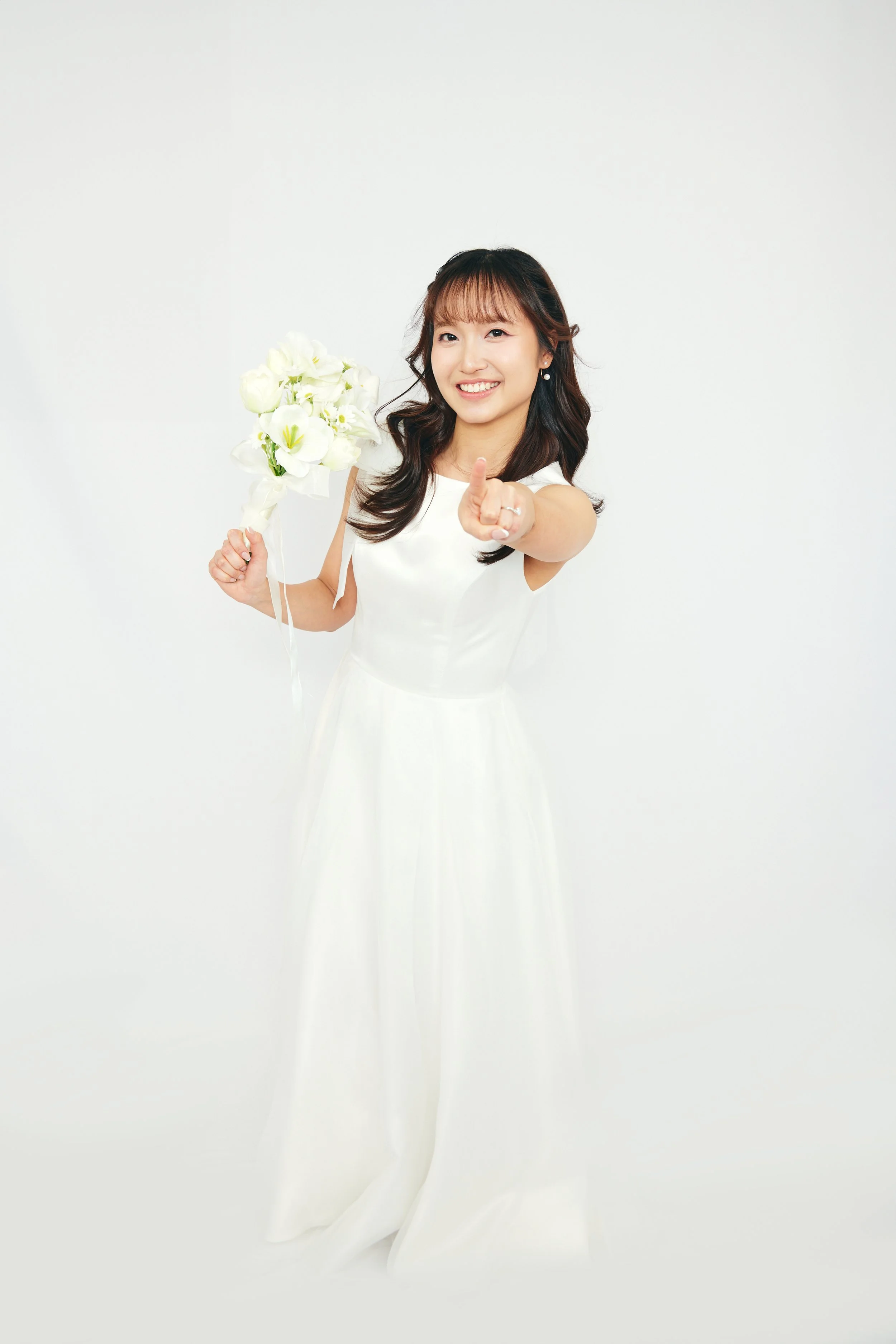 Young woman in white wedding dress holding bouquet of white flowers, smiling and pointing towards the camera.