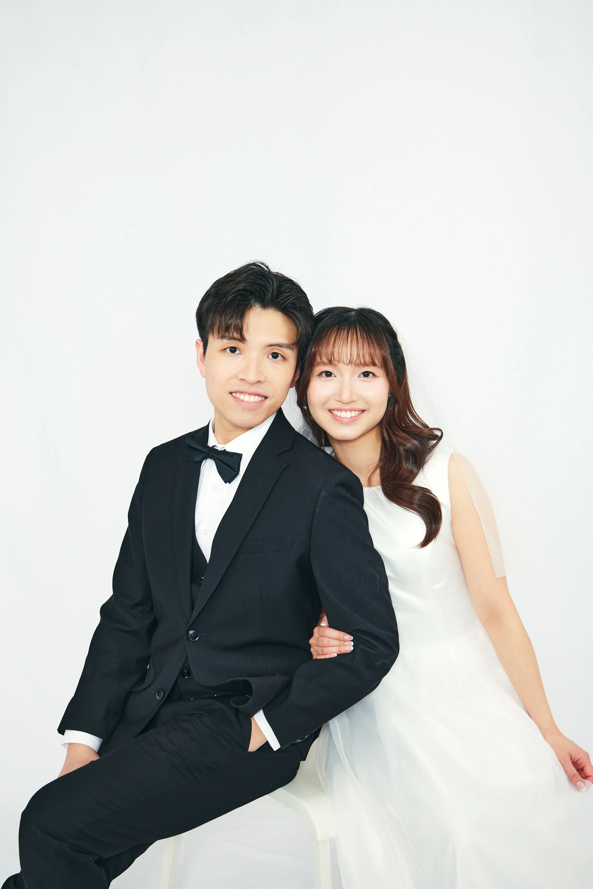 A young couple dressed in formal wedding attire, sitting close together and smiling at the camera, with a plain white background.