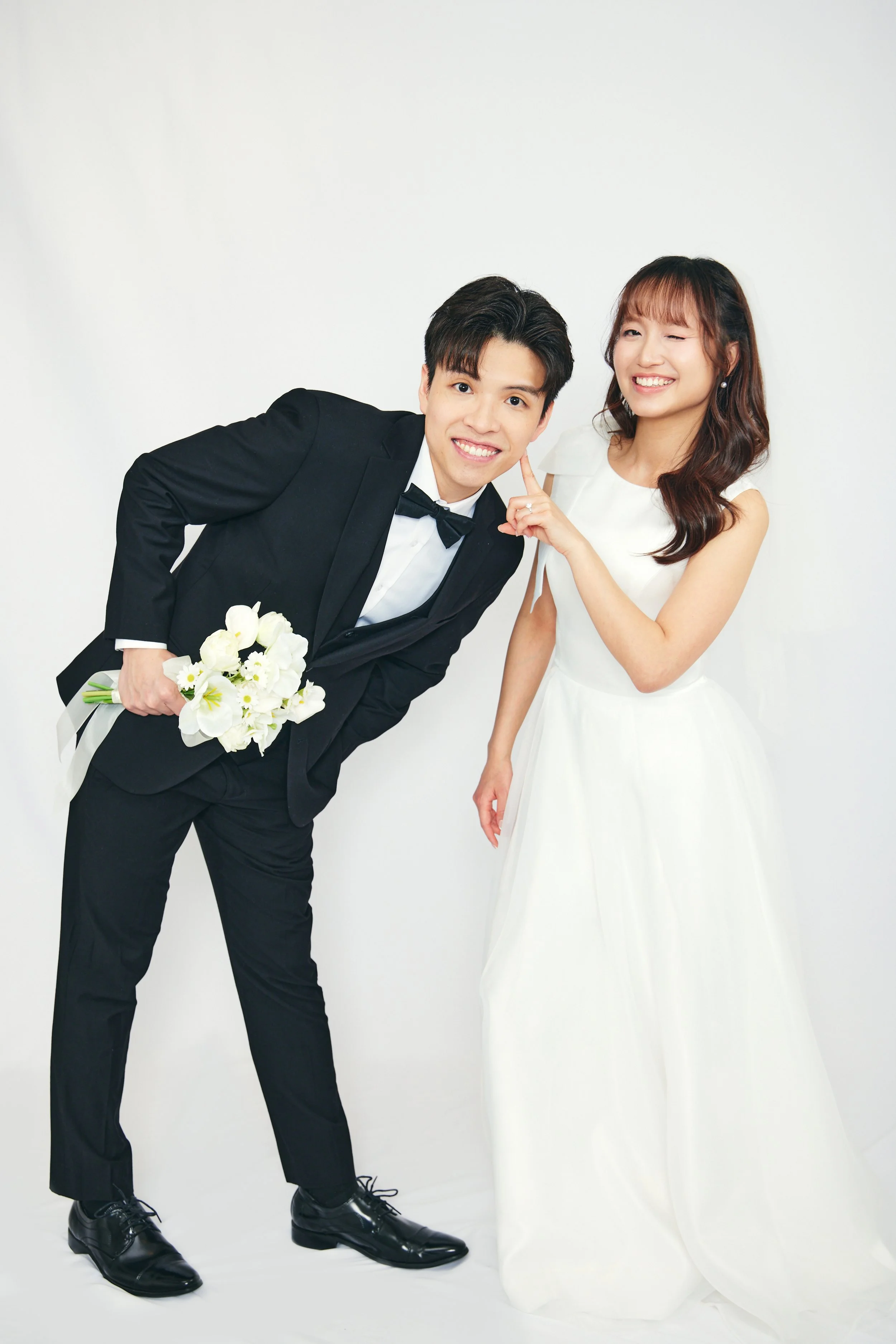 A couple dressed in wedding attire, the groom in a black tuxedo holding a bouquet of white flowers, and the bride in a white wedding gown, standing together against a plain white background.