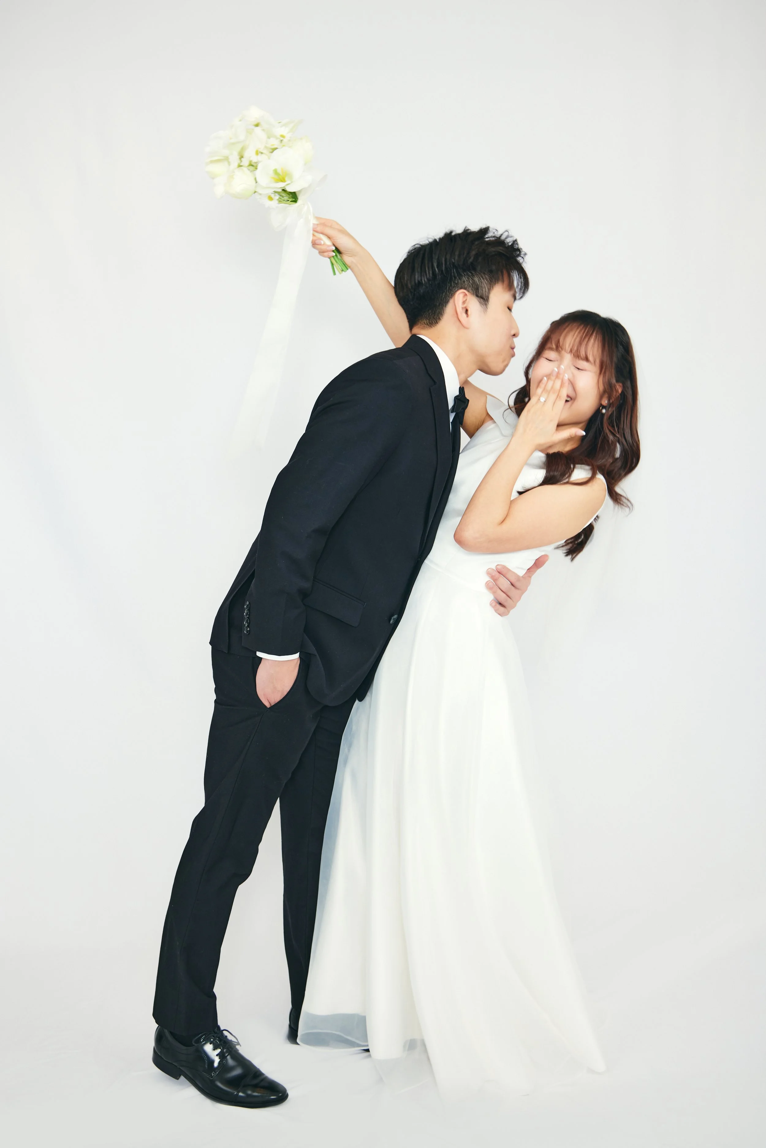 A bride and groom share a playful moment, with the groom kissing the bride on the cheek as she laughs and covers her mouth. The groom is in a black suit and the bride in a white wedding dress, against a plain white background, with a bouquet of white