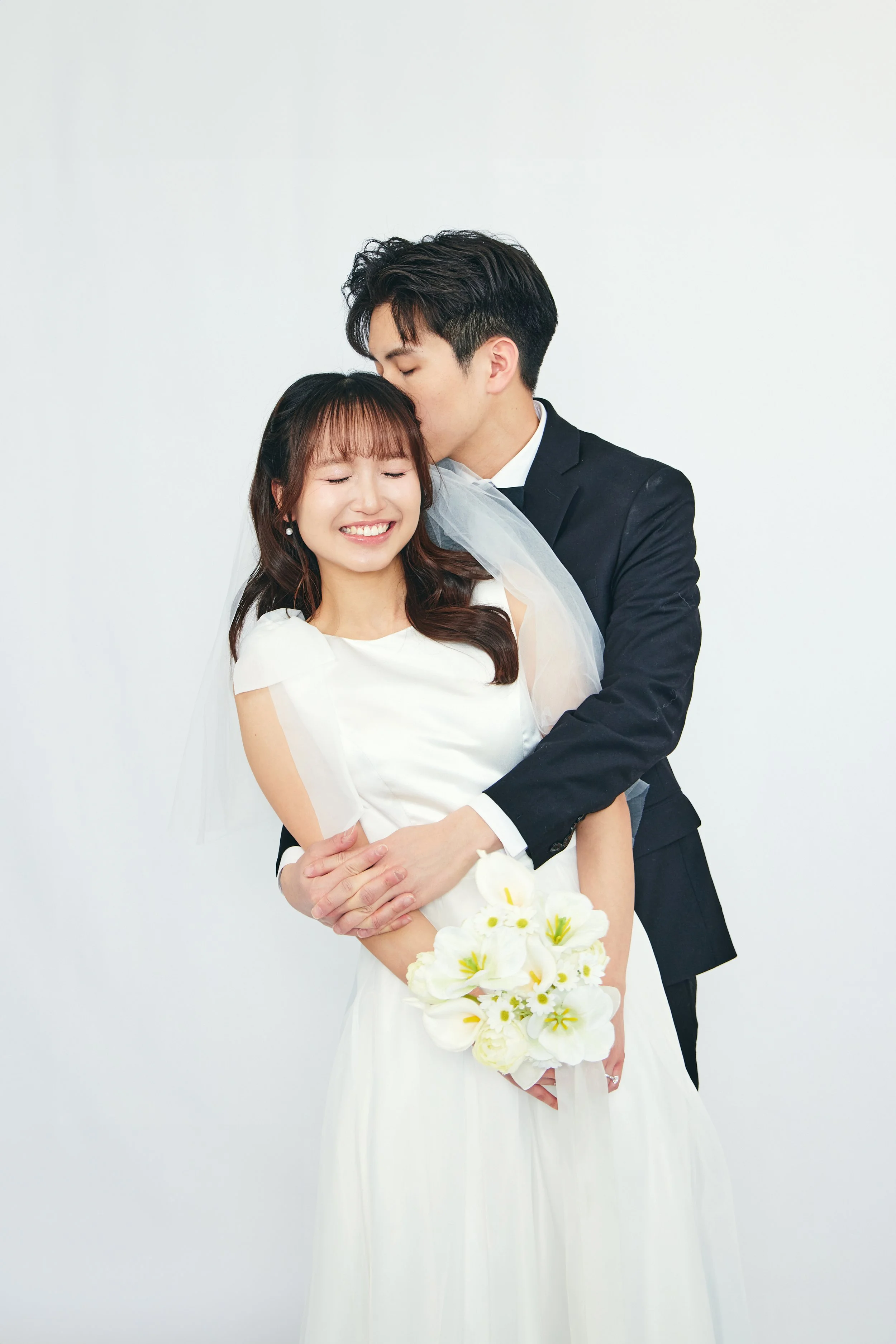 A bride and groom in wedding attire embracing each other against a white background, with the groom kissing the bride's forehead. The bride holds a bouquet of white flowers and is smiling with eyes closed.