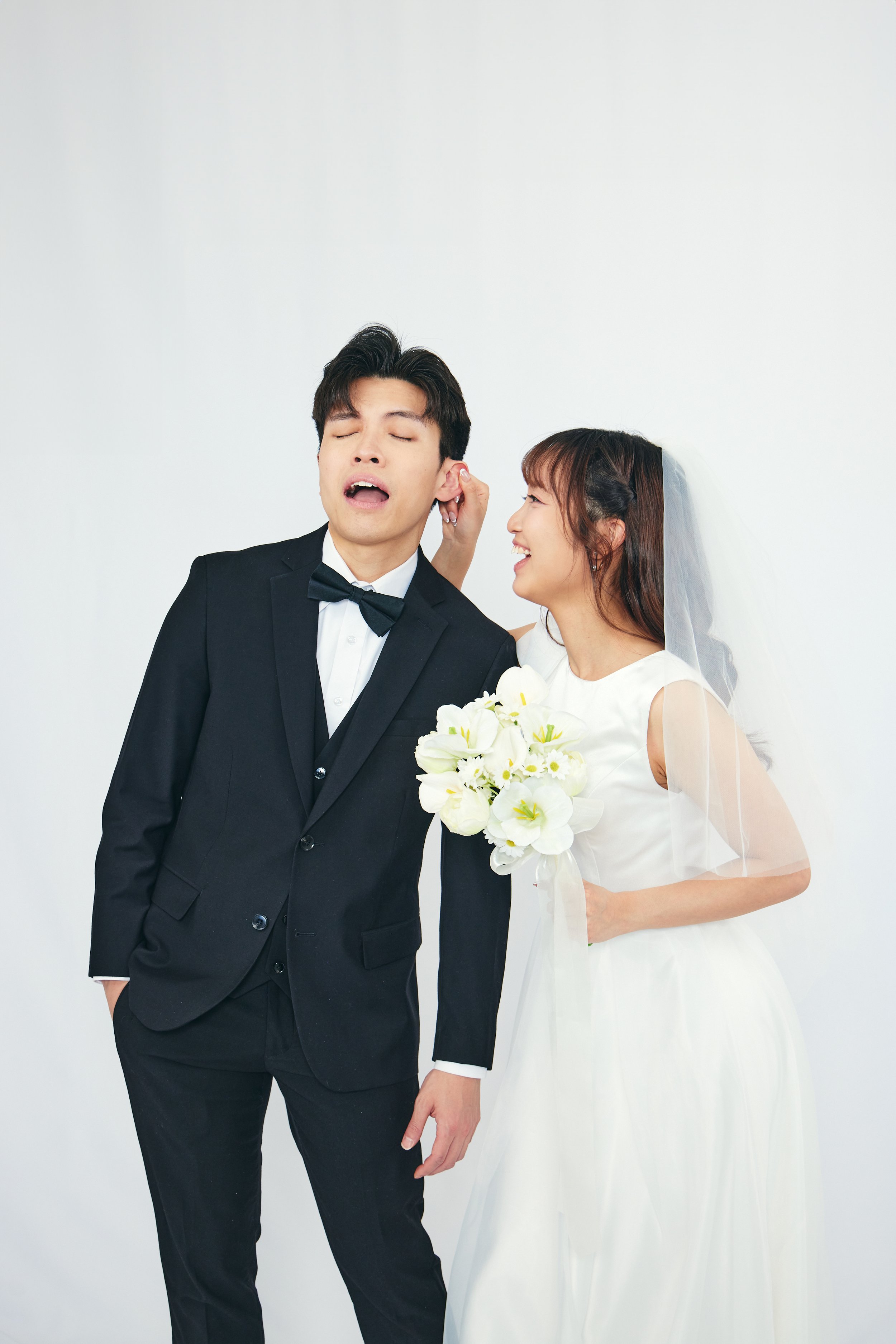 Bride in a white wedding dress holding a bouquet of white flowers, leaning towards groom in a black suit, white shirt, and black bow tie, who has his eyes closed with a playful expression.
