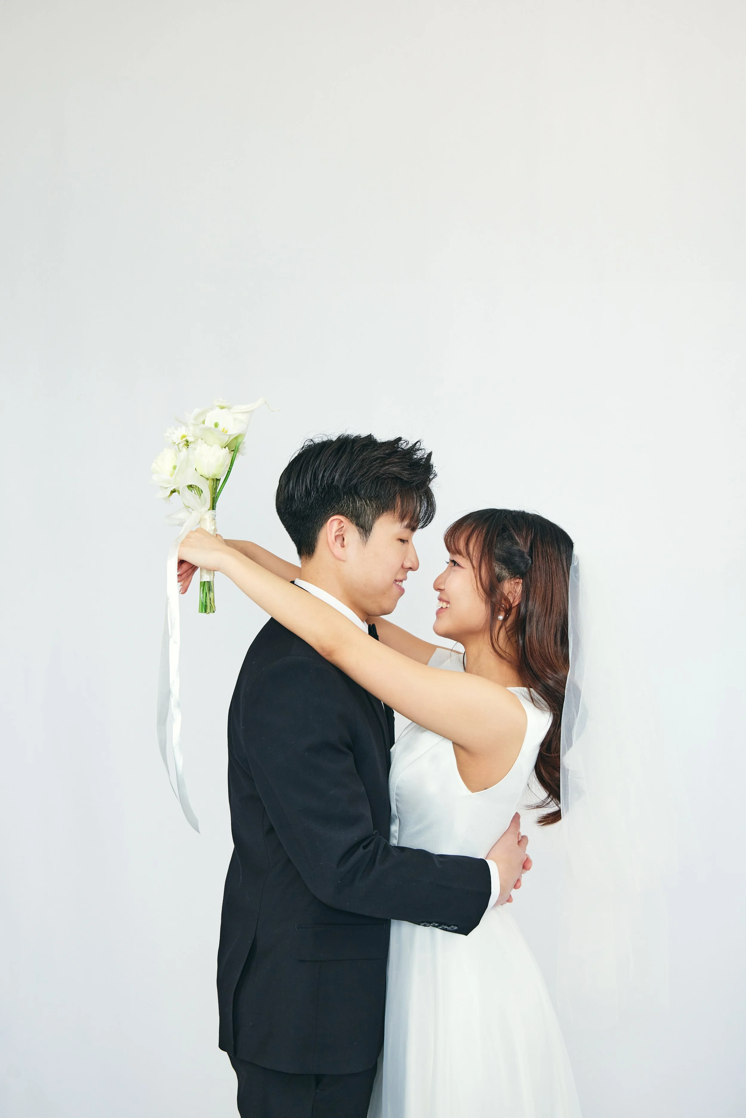 A newlywed Asian couple stands close, smiling at each other. The woman has her arms around the man's neck, holding a bouquet of white calla lilies, while the man gently holds her waist. The background is plain white.