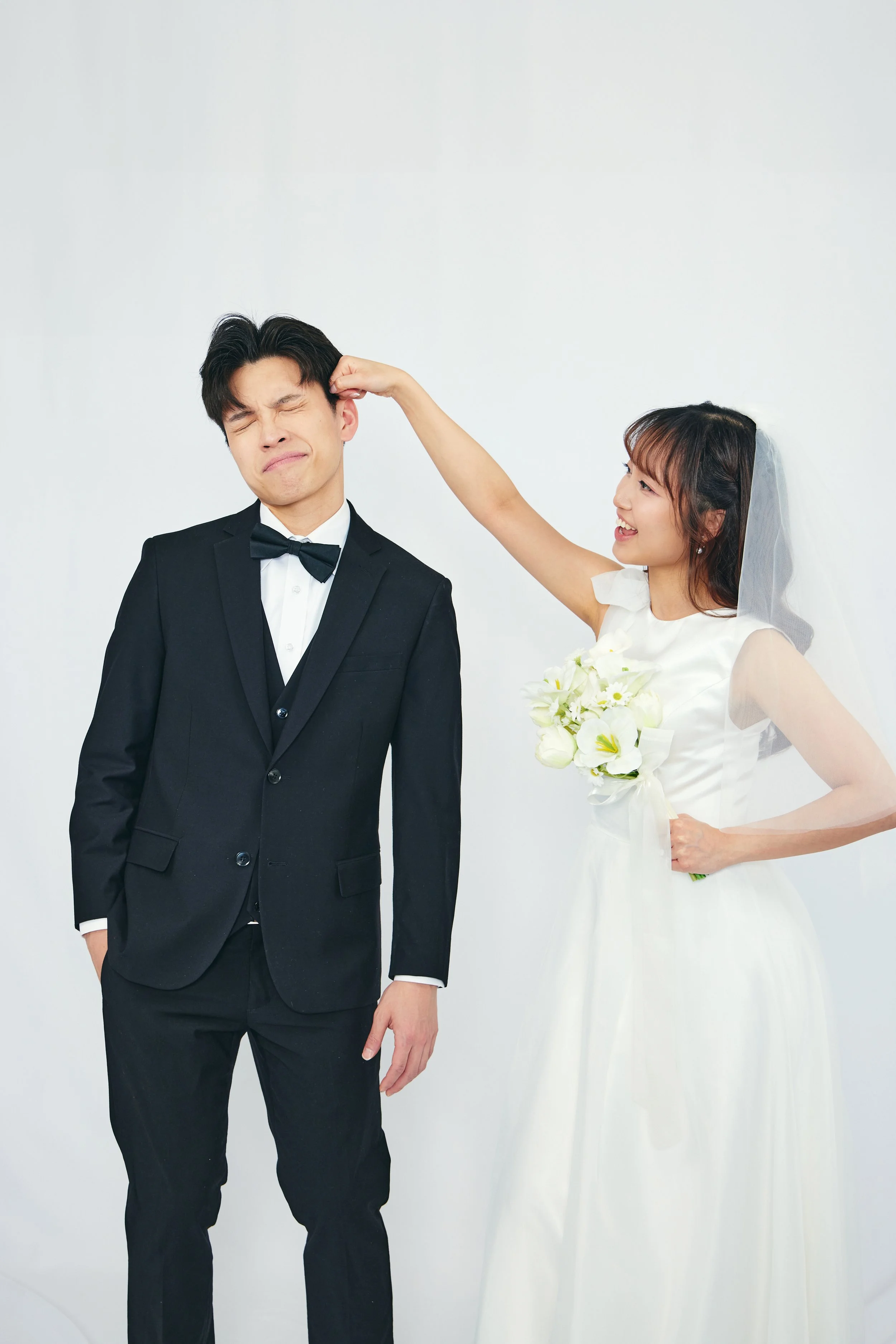 A bride in a white wedding dress holding a bouquet, playfully hitting a groom in a black tuxedo on the head, both standing against a plain white background.