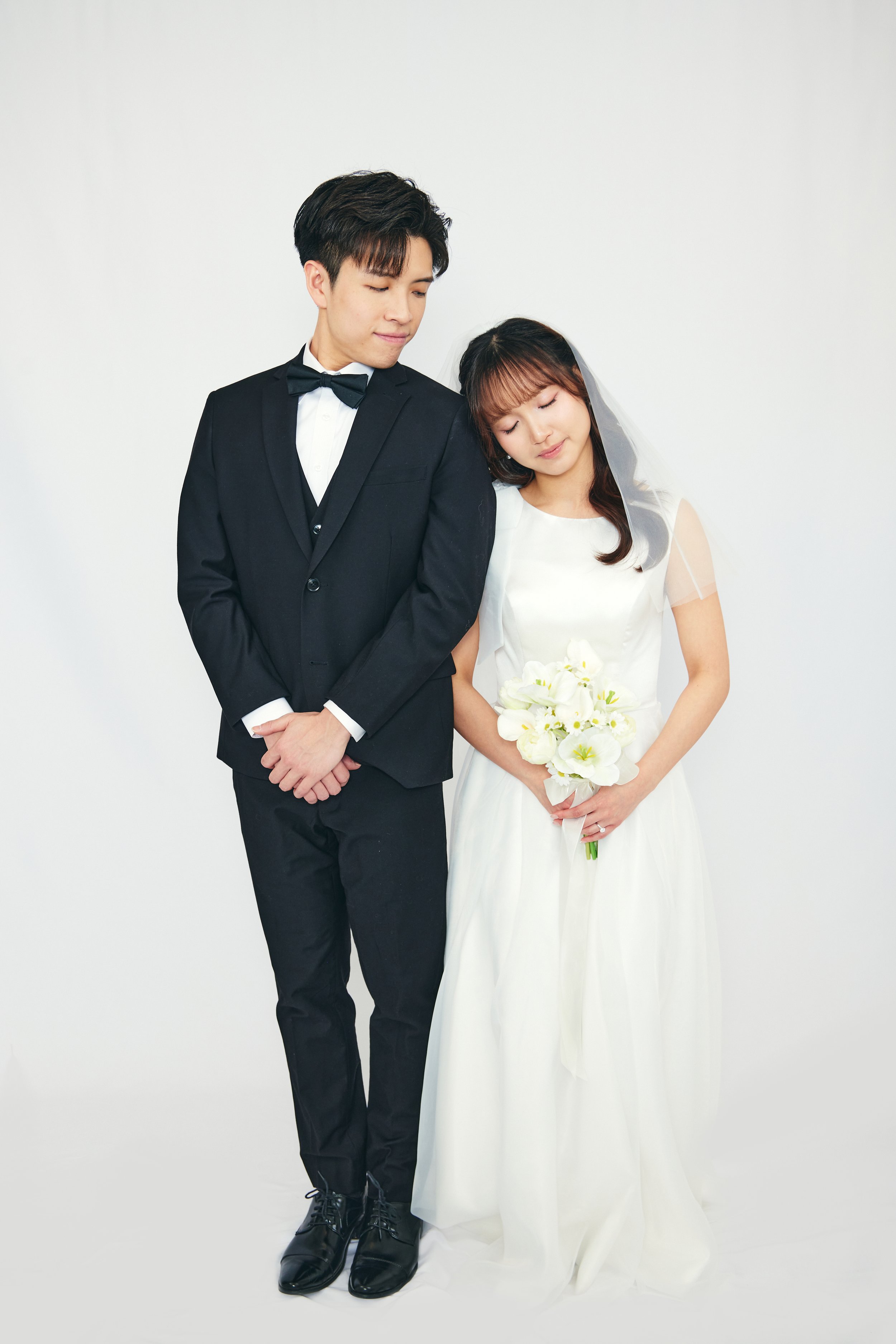 A bride in a white wedding dress holding a bouquet of white flowers stands next to a groom in a black suit and bow tie, both with their eyes closed and head slightly tilted towards each other, against a plain white background.