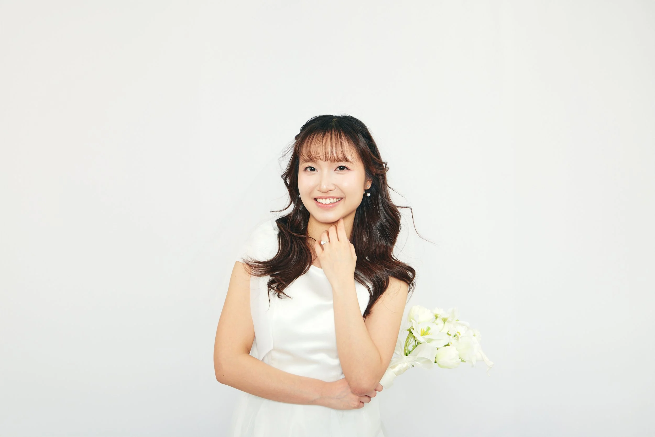 A young woman in a white dress holding a bouquet of white flowers, smiling, with long wavy brown hair and bangs, standing against a plain white background.