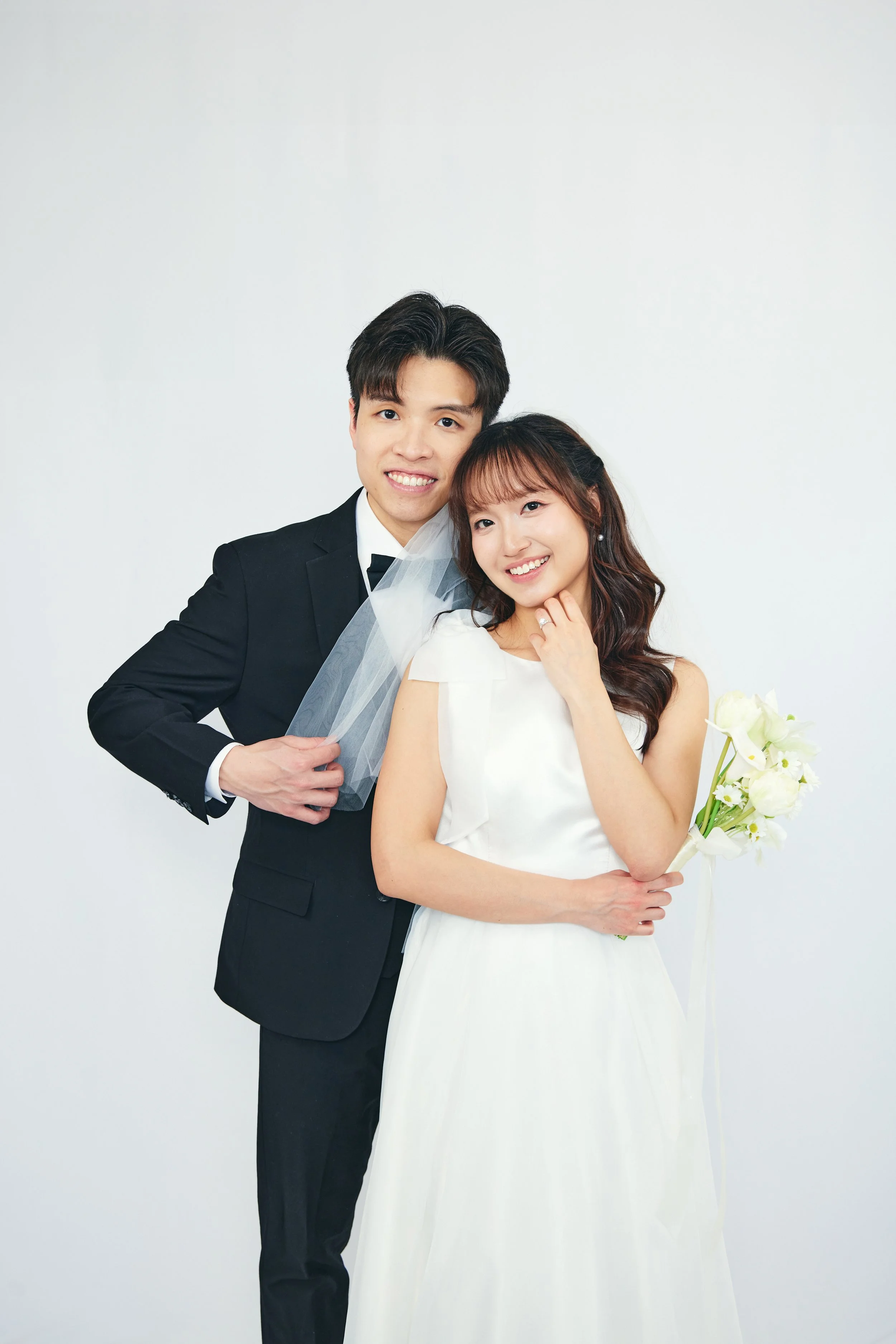 A young couple on their wedding day, the groom in a black tuxedo and the bride in a white wedding dress holding a bouquet of white flowers, smiling against a plain white background.