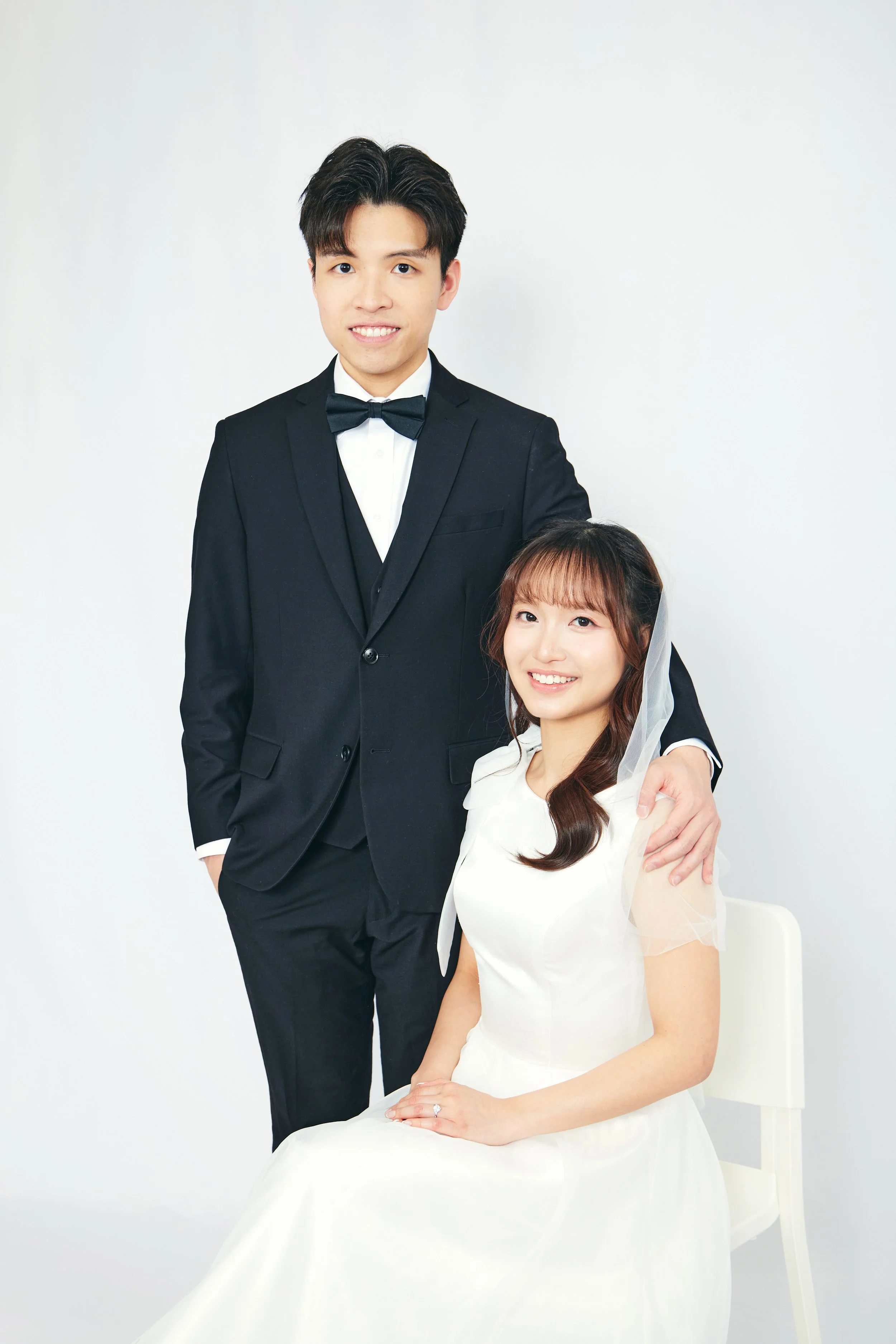 A young man in a tuxedo and a young woman in a wedding dress pose together against a plain white background.