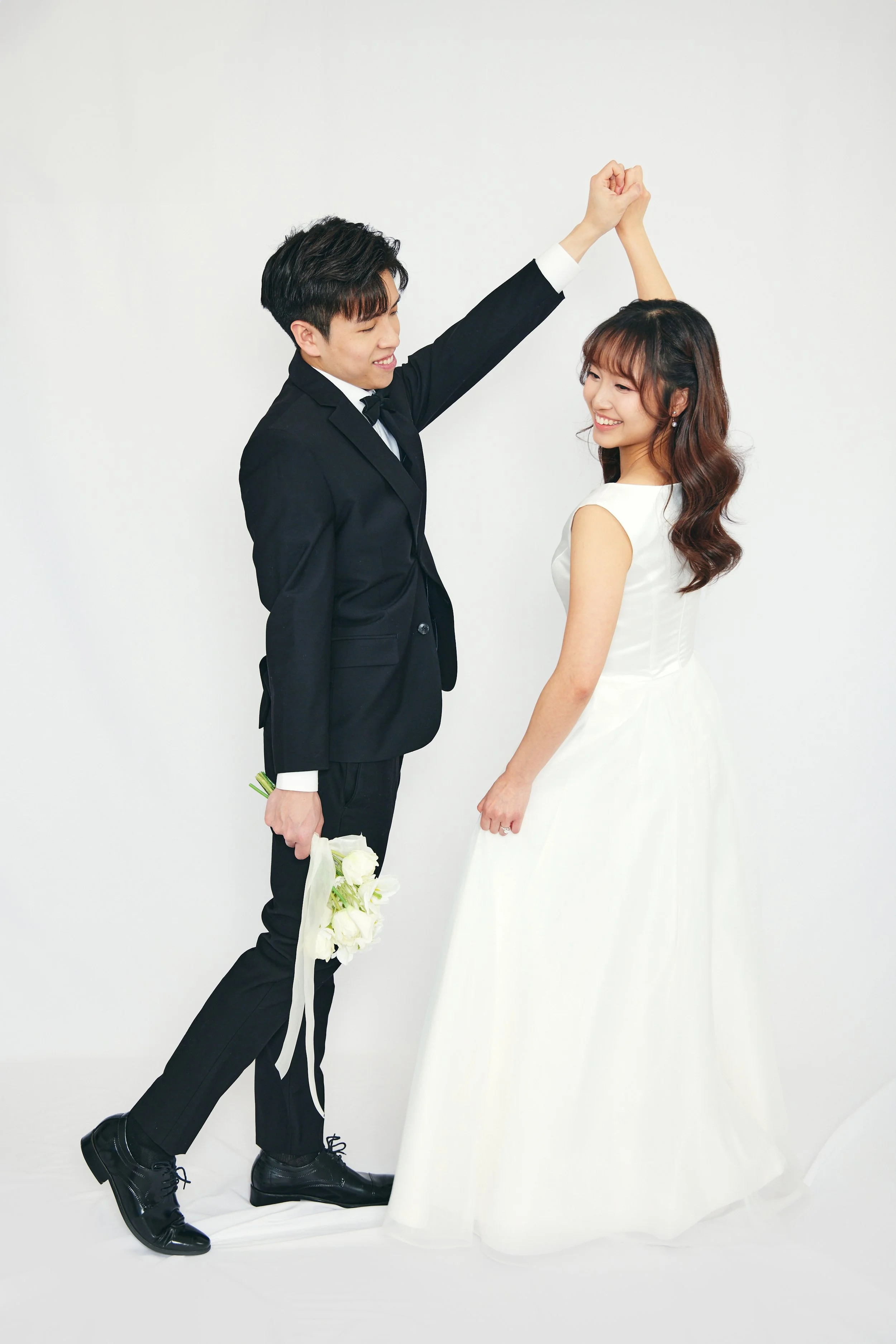A couple dressed in wedding attire, with the groom in a black suit holding a bouquet and the bride in a white wedding dress, smiling at each other against a plain white background.