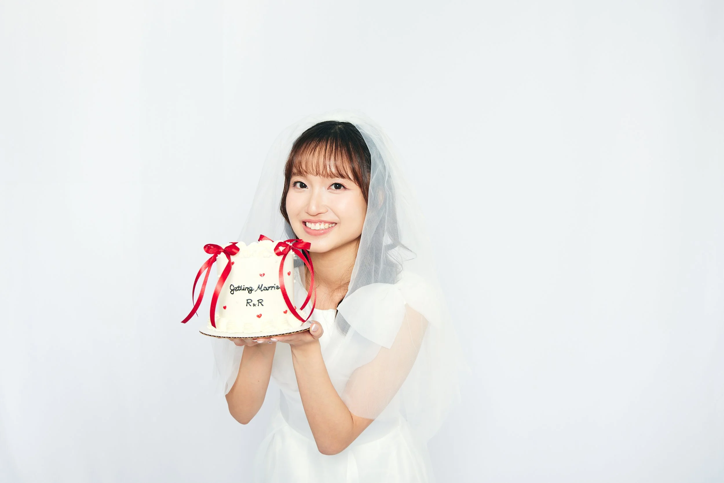 Young woman wearing a wedding dress and veil holding a cake decorated with red ribbons and bows, with writing that says 'Getting Married R & R', smiling against a plain white background.