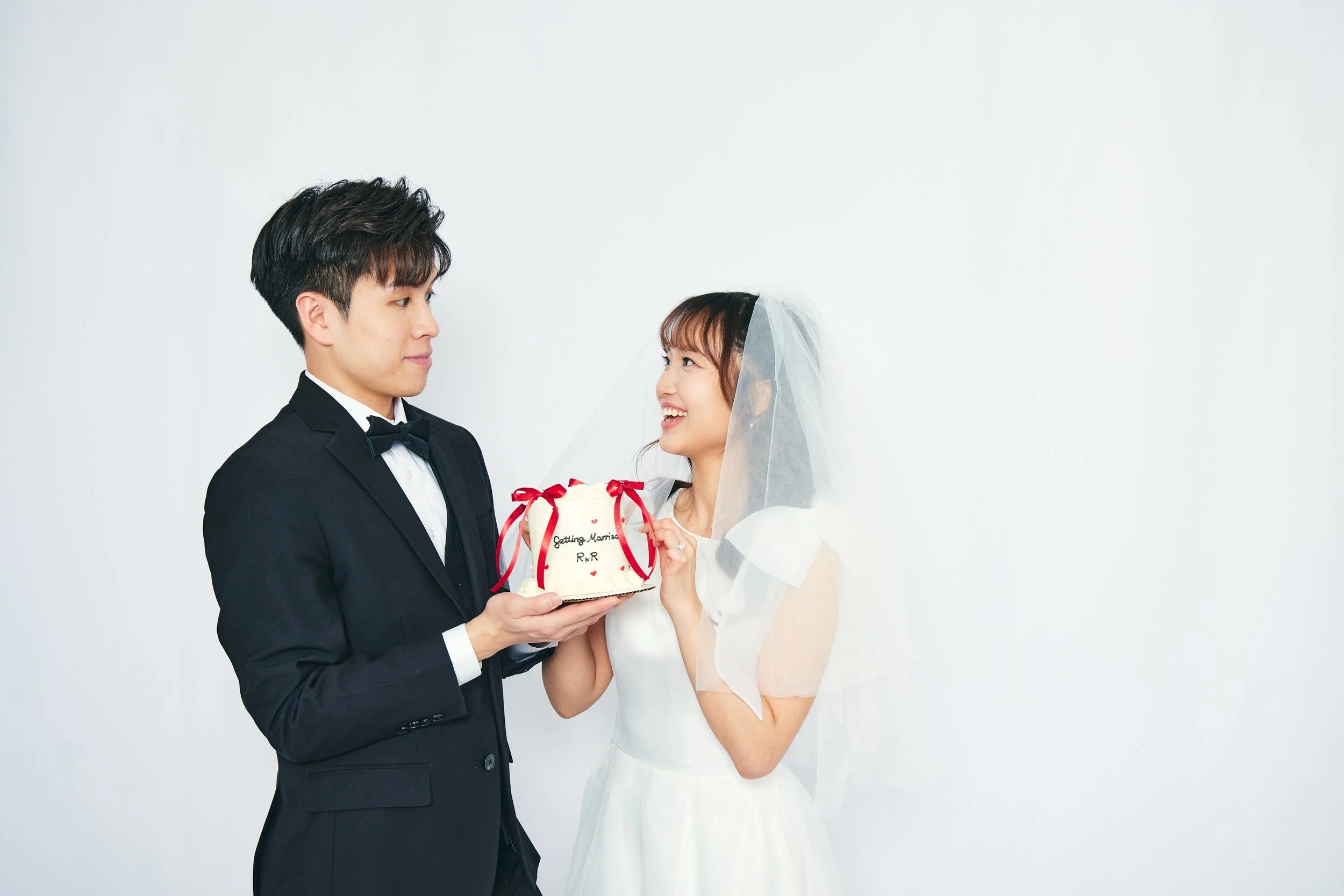 A groom in a black tuxedo and bow tie holding a wedding cake with a bride in a white wedding dress and veil, smiling at each other against a plain white background.