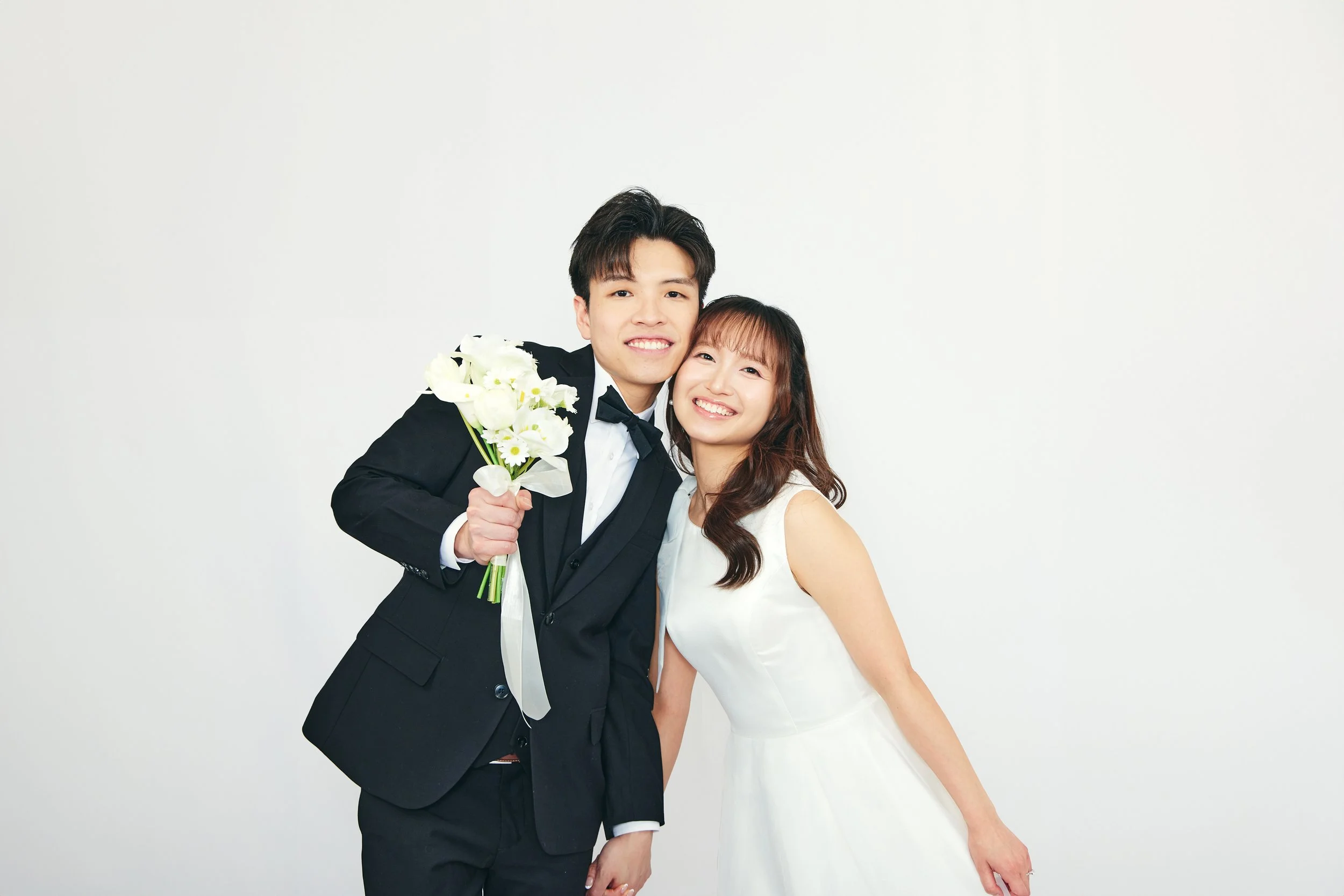 A happy couple dressed in wedding attire, the man in a black tuxedo holding a bouquet of white flowers, smiling against a plain white background.